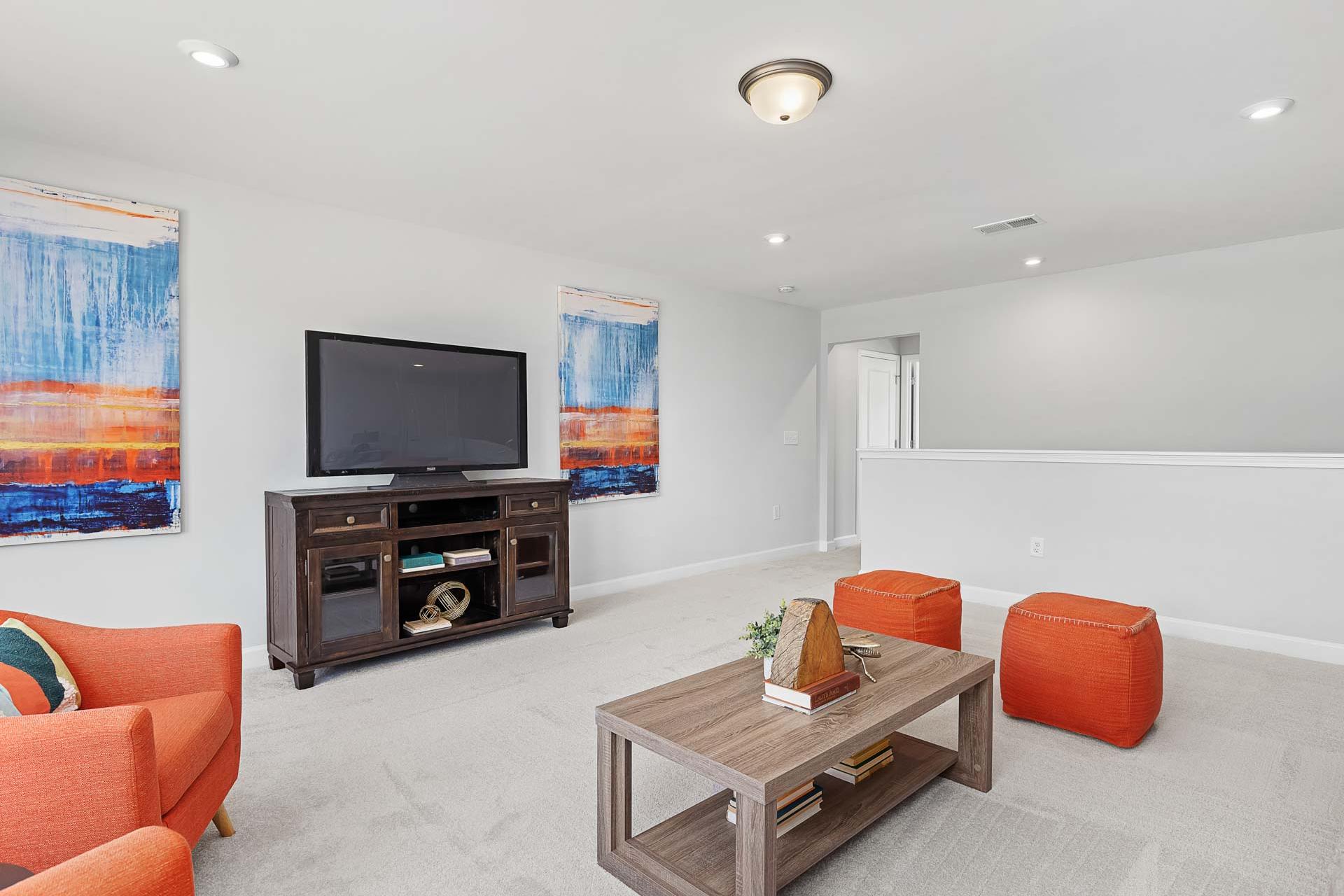 Cozy loft bonus room in Glenmere Knightdale NC with wooden TV entertainment center, abstract blue-orange wall art, orange pouf seating, and modern coffee table
