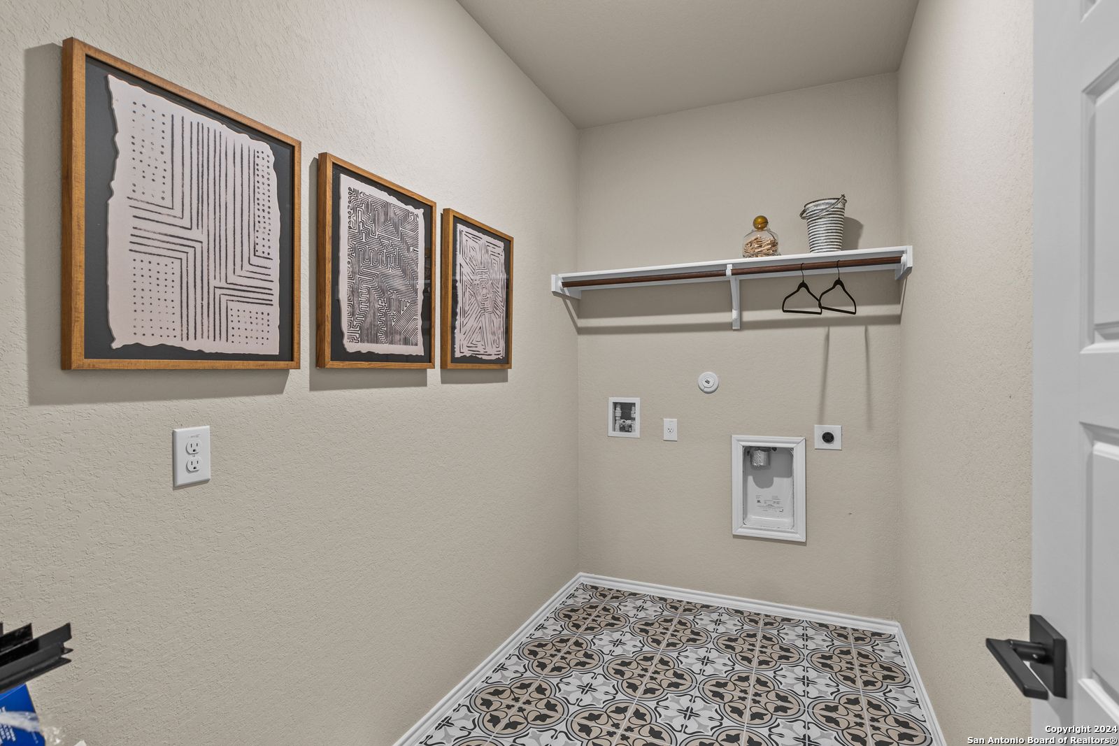 Modern laundry room with abstract framed art, utility shelf, washer/dryer hookups, and geometric tile floor in Davidson Homes Douglas F, San Antonio