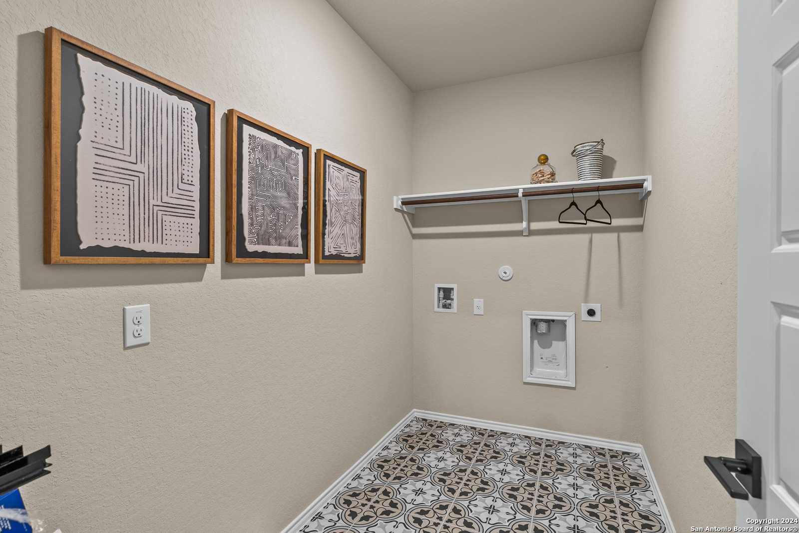 Modern laundry room with abstract framed art, utility shelf, washer/dryer hookups, and geometric tile floor in Davidson Homes Douglas F, San Antonio