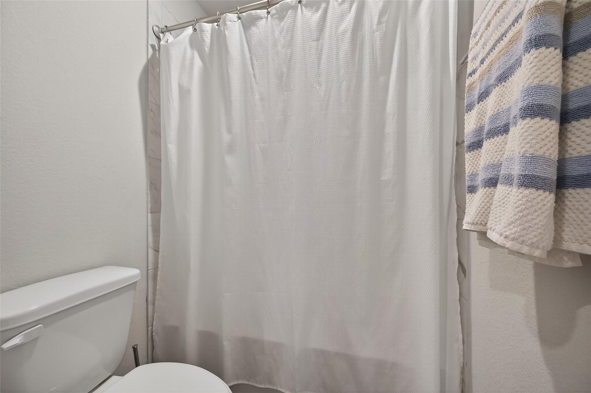 Bright bathroom with white shower curtain, white toilet, and blue striped towel in Davidson Homes The Brazos E, Magnolia, Texas