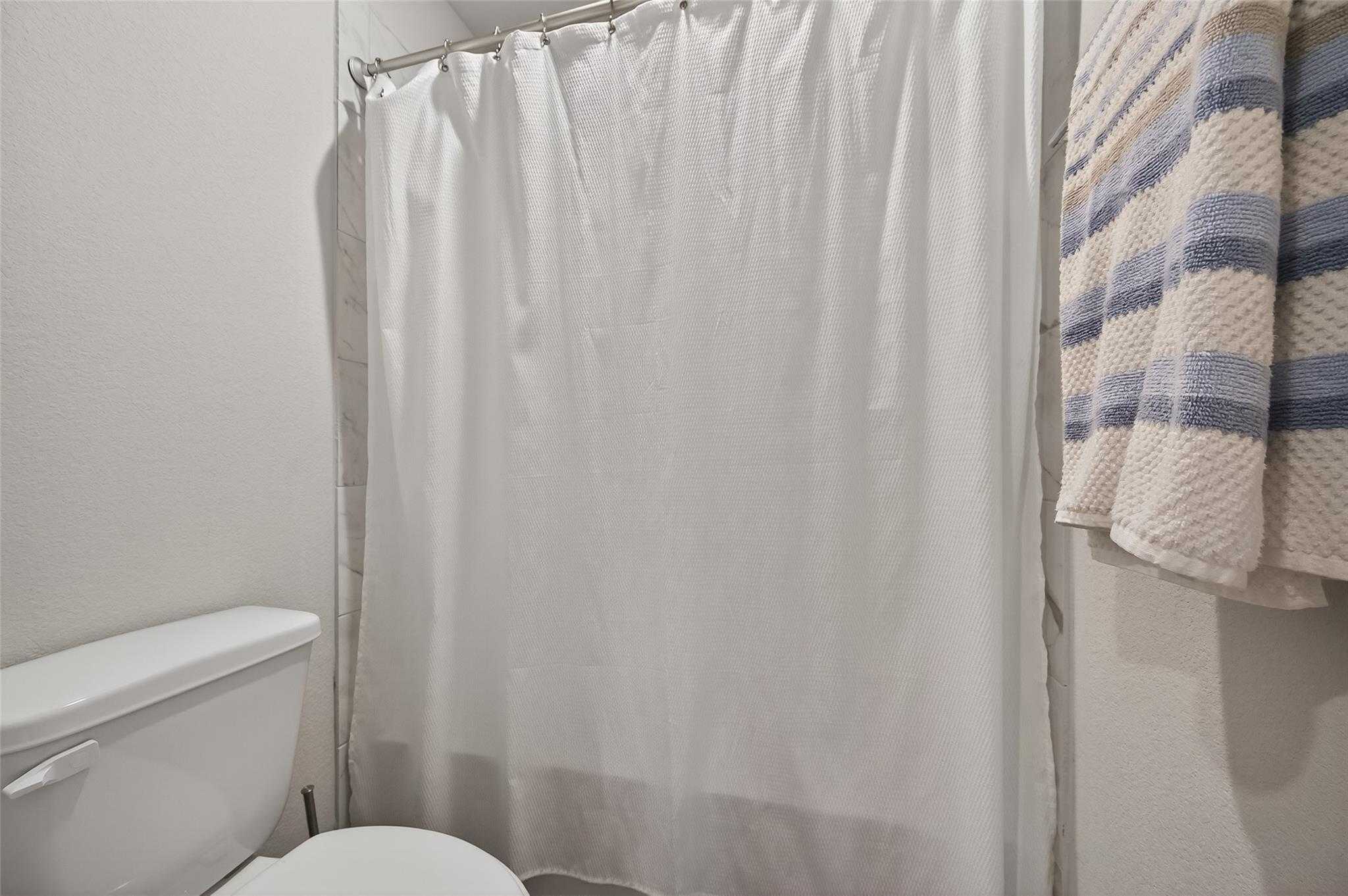 Bright bathroom with white shower curtain, white toilet, and blue striped towel in Davidson Homes The Brazos E, Magnolia, Texas