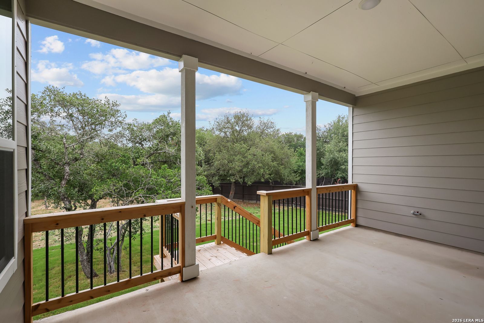 Covered porch with white columns, wooden railing, and steps to lush green backyard in Davidson Homes The Belmont H, Castroville, Texas