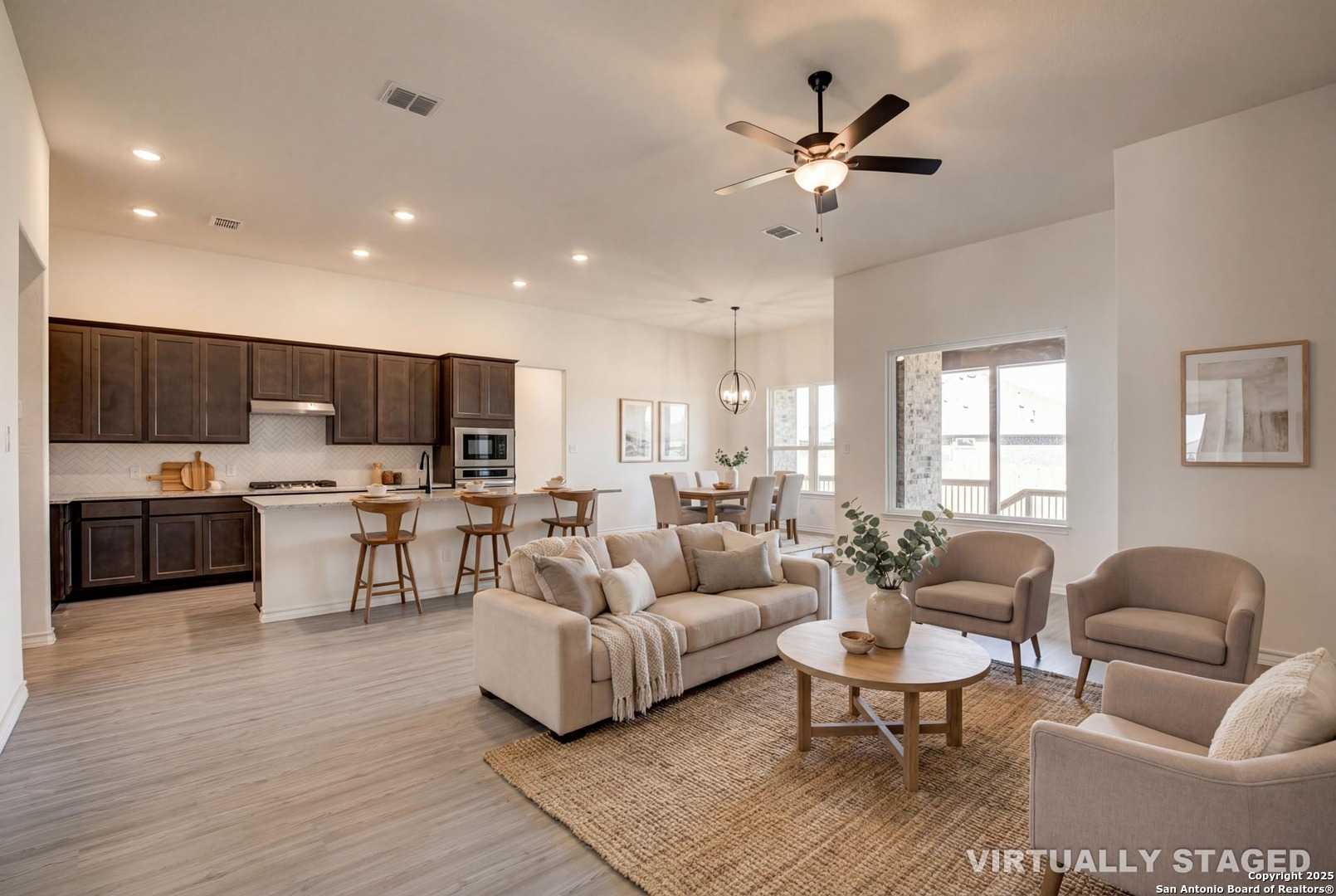 Open-concept living and kitchen with beige sofa, dark cabinets, island bar, and dining area in The Garner B, Castroville, Texas