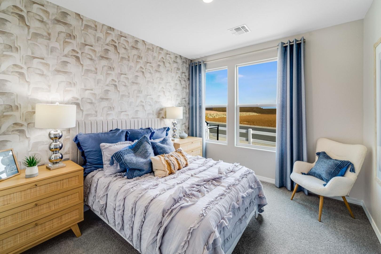 Spacious master bedroom in The Newport C showcasing tufted gray bed, blue pillows, modern wallpaper, and scenic view through large windows