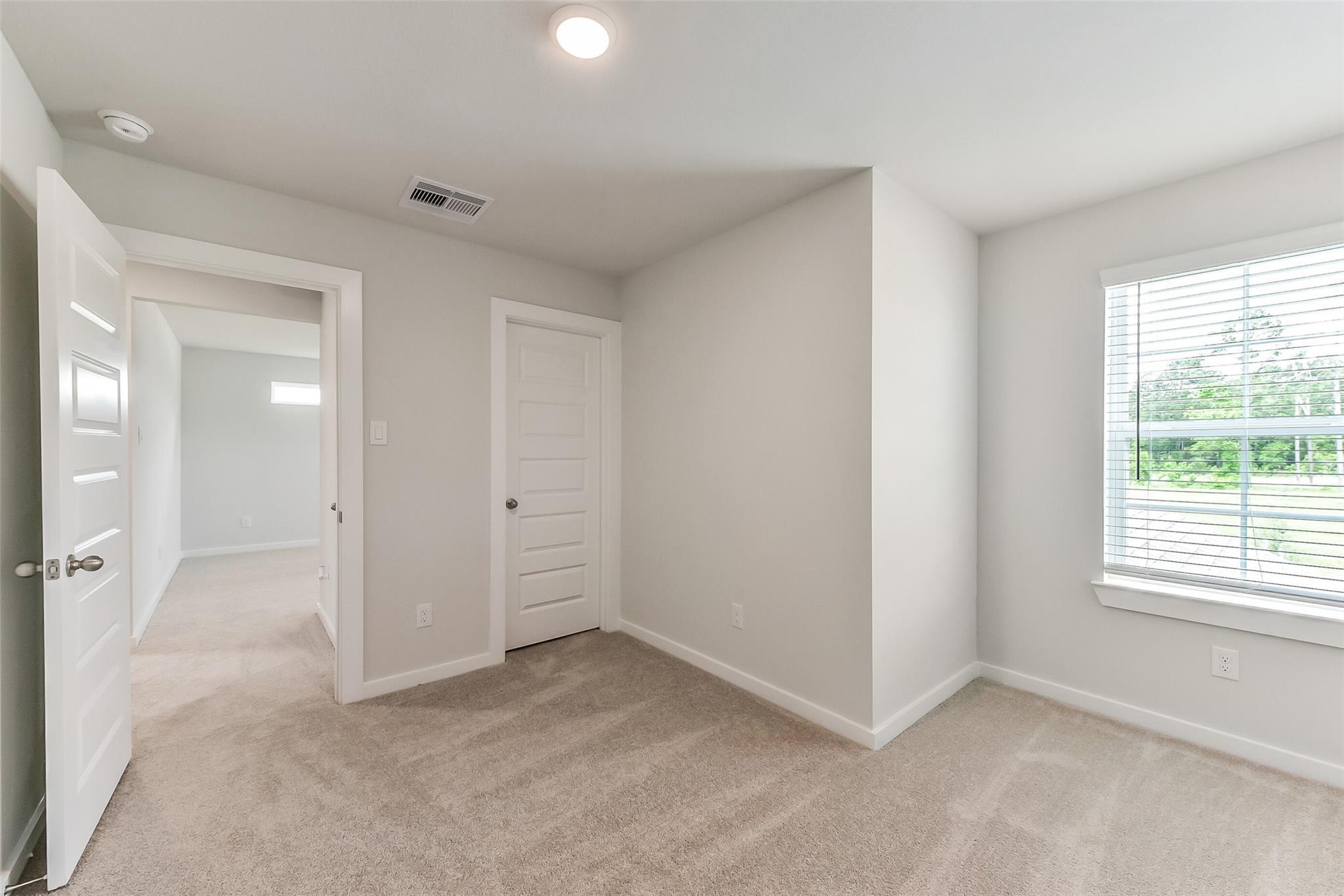 Bright secondary bedroom with neutral walls, beige carpet, large window with blinds overlooking trees, closet door in Davidson Homes The Sabine F, Dayton Texas