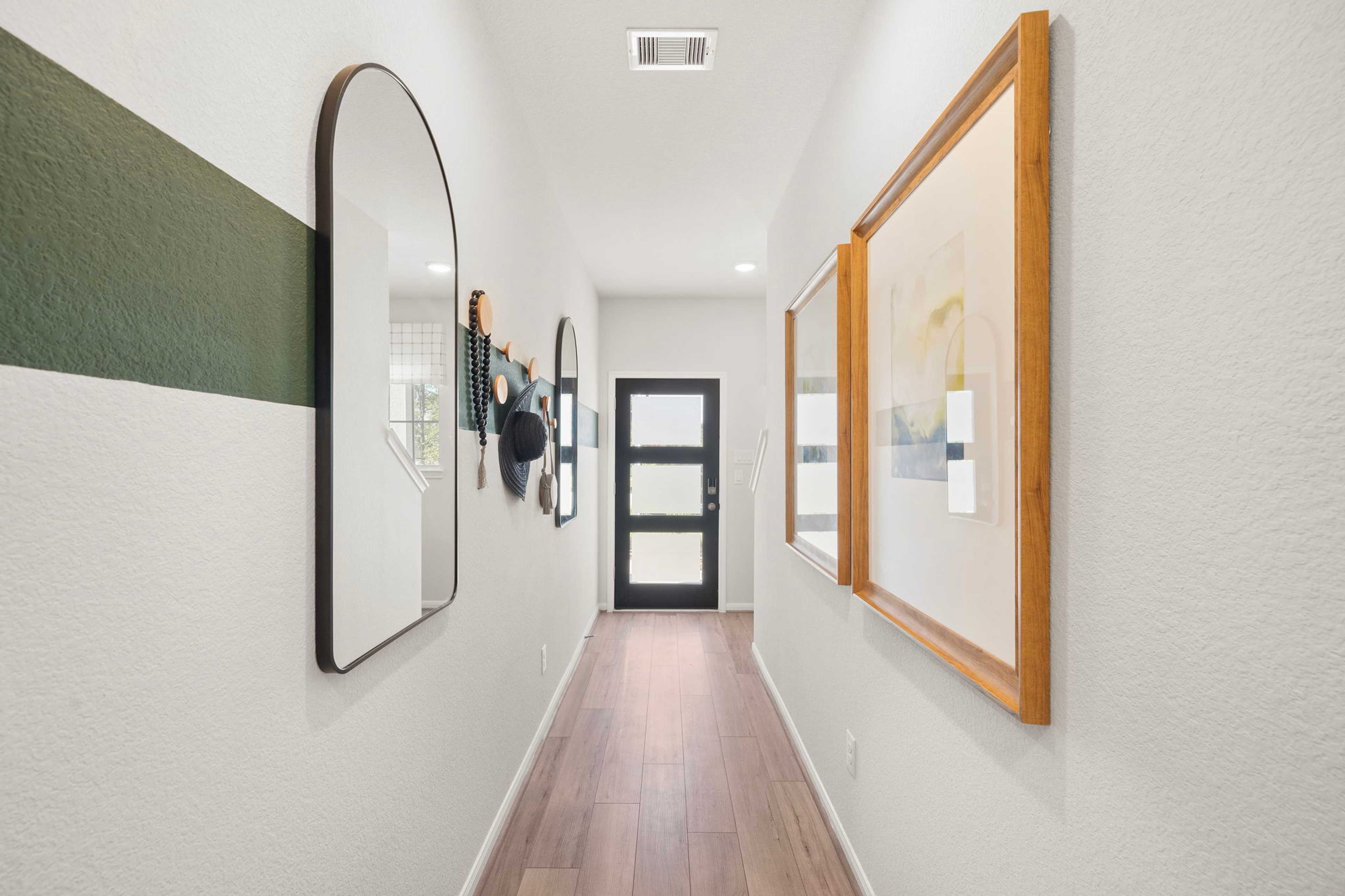 Spacious hallway at Caney Creek Place in Conroe Texas with hardwood floors, arched mirror, wall art, hats and black front door