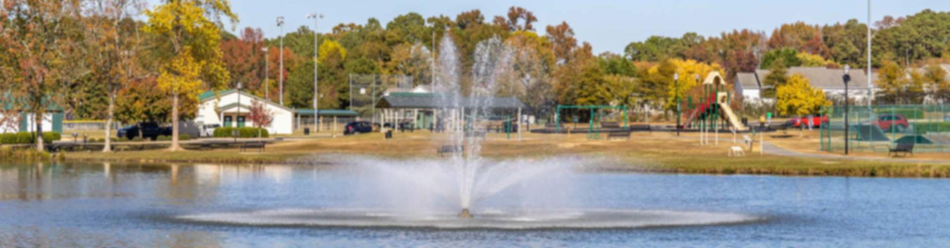 Scenic fountain spraying in pond at Angier community park with autumn trees, playground, and sports fields