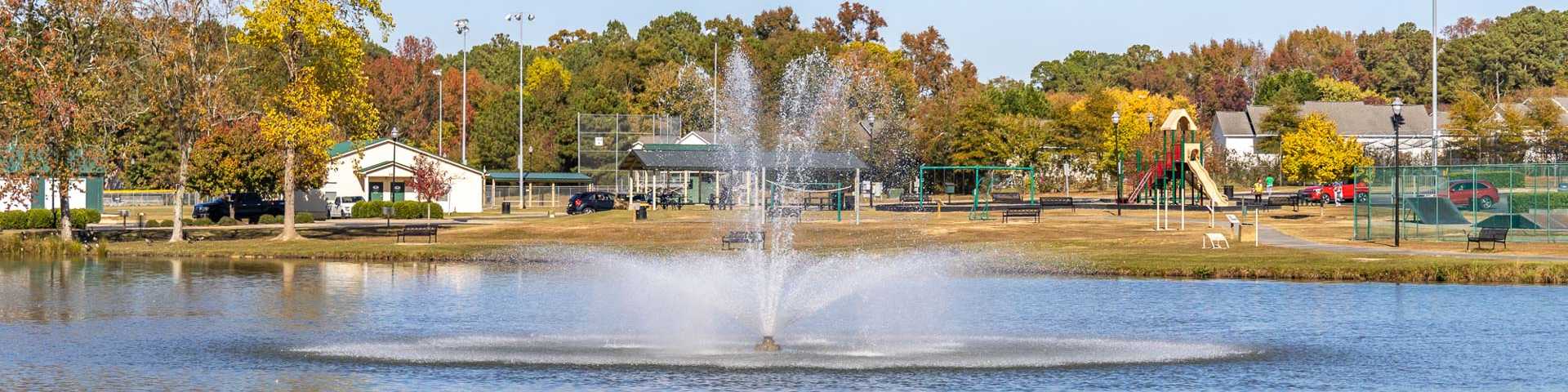 Scenic fountain spraying in pond at Angier community park with autumn trees, playground, and sports fields