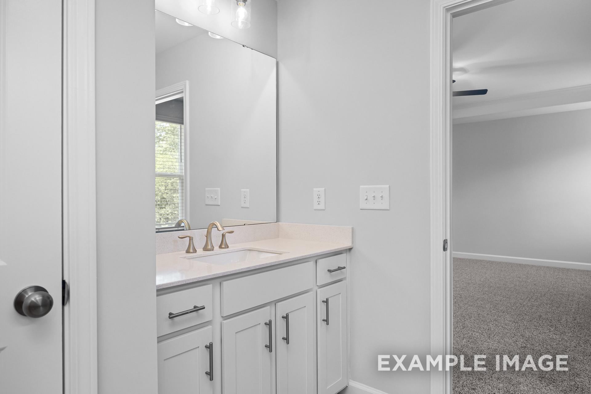 Master bathroom in The Preston B with double vanity, gold faucet, white shaker cabinets, and open door to carpeted bedroom