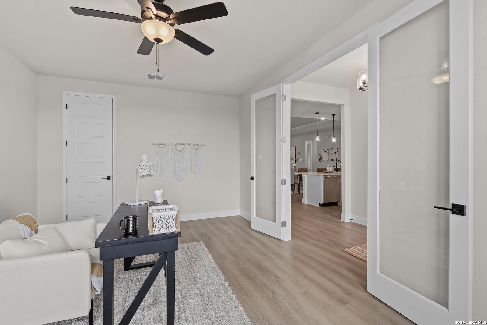 Cozy home office with black desk, white armchair, woven decor, and frosted French doors to kitchen in Davidson Homes The Lanier G, Castroville TX