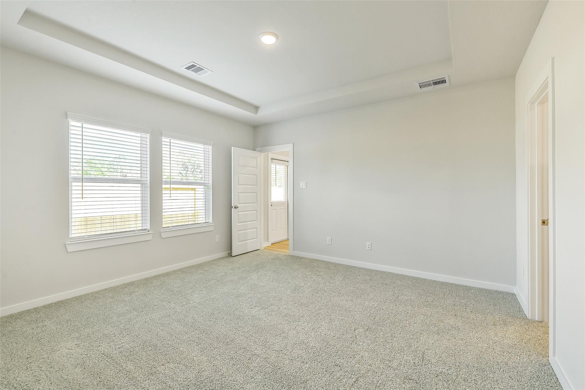 Spacious empty bedroom with beige carpet, light gray walls, window blinds, and open bathroom door in Davidson Homes The San Marcos E, Conroe, Texas