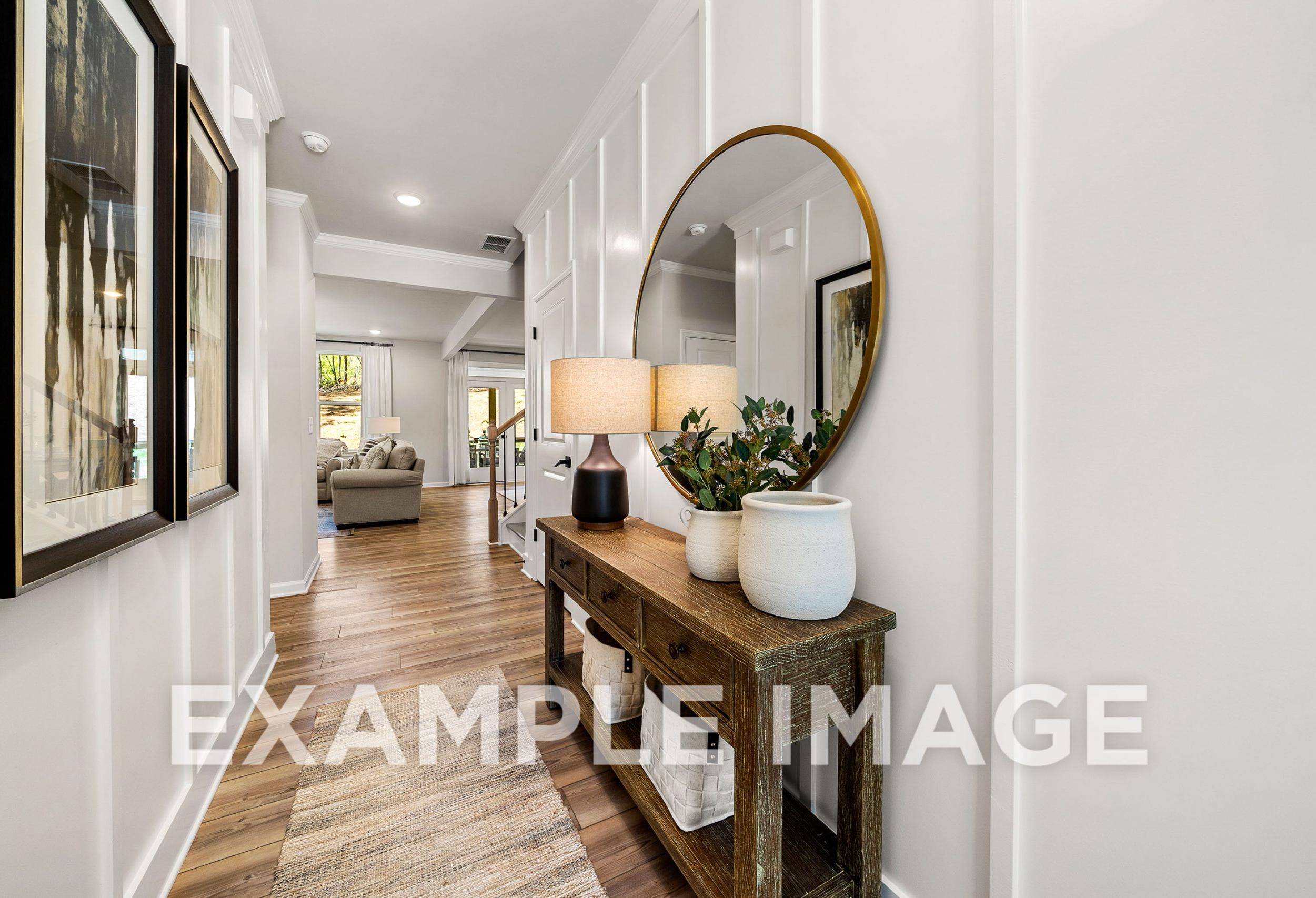 Spacious hallway in The Hickory B home with white wainscoting, wooden console table, gold mirror, lamps, and plants leading to open living area