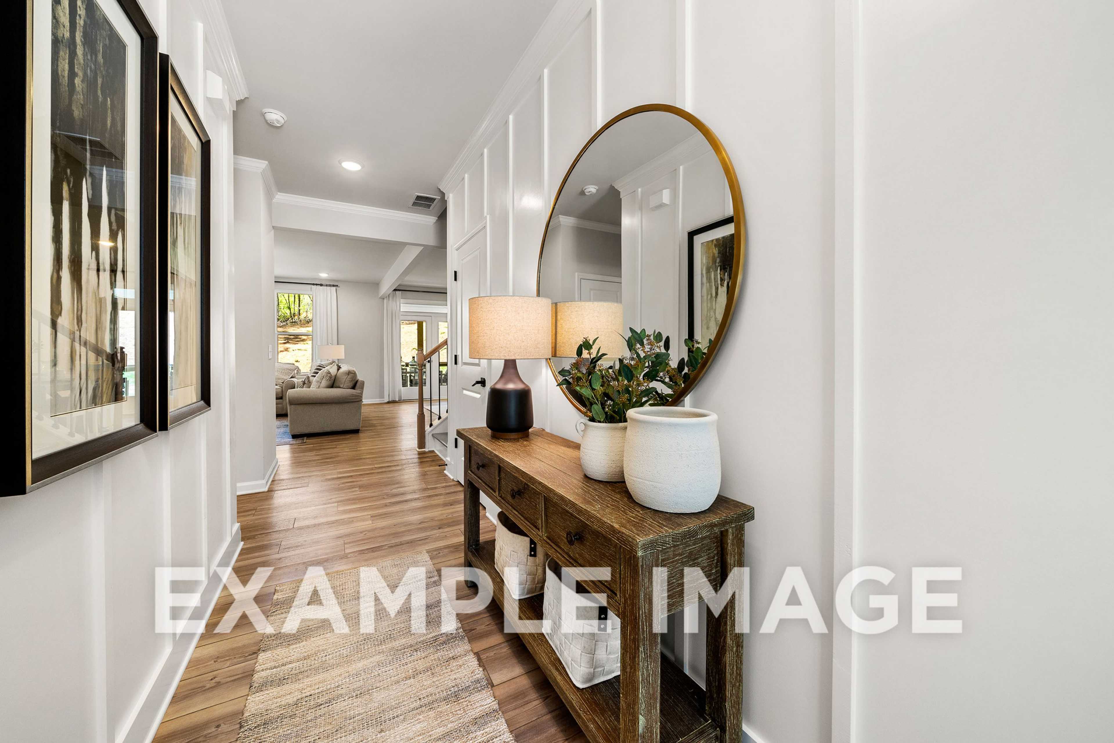 Spacious hallway in The Hickory home with white wainscoting, round gold mirror, wooden console table, lamps, plants, and artwork