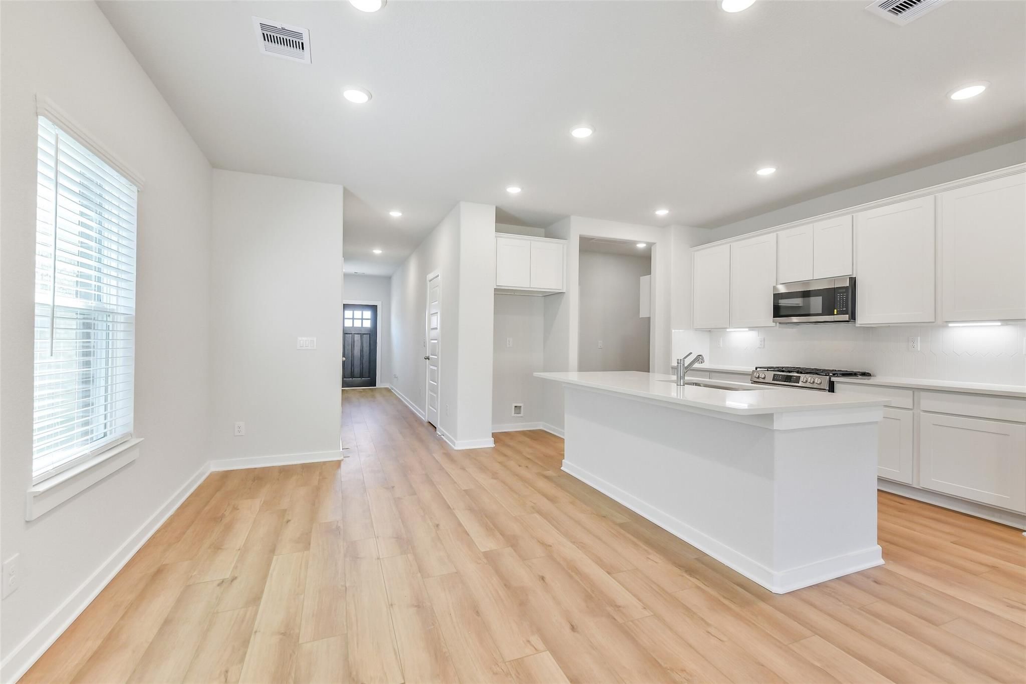 Open-concept white kitchen with large island, stainless appliances, hardwood floors, and hallway view in Davidson Homes The Blanco E, Magnolia TX