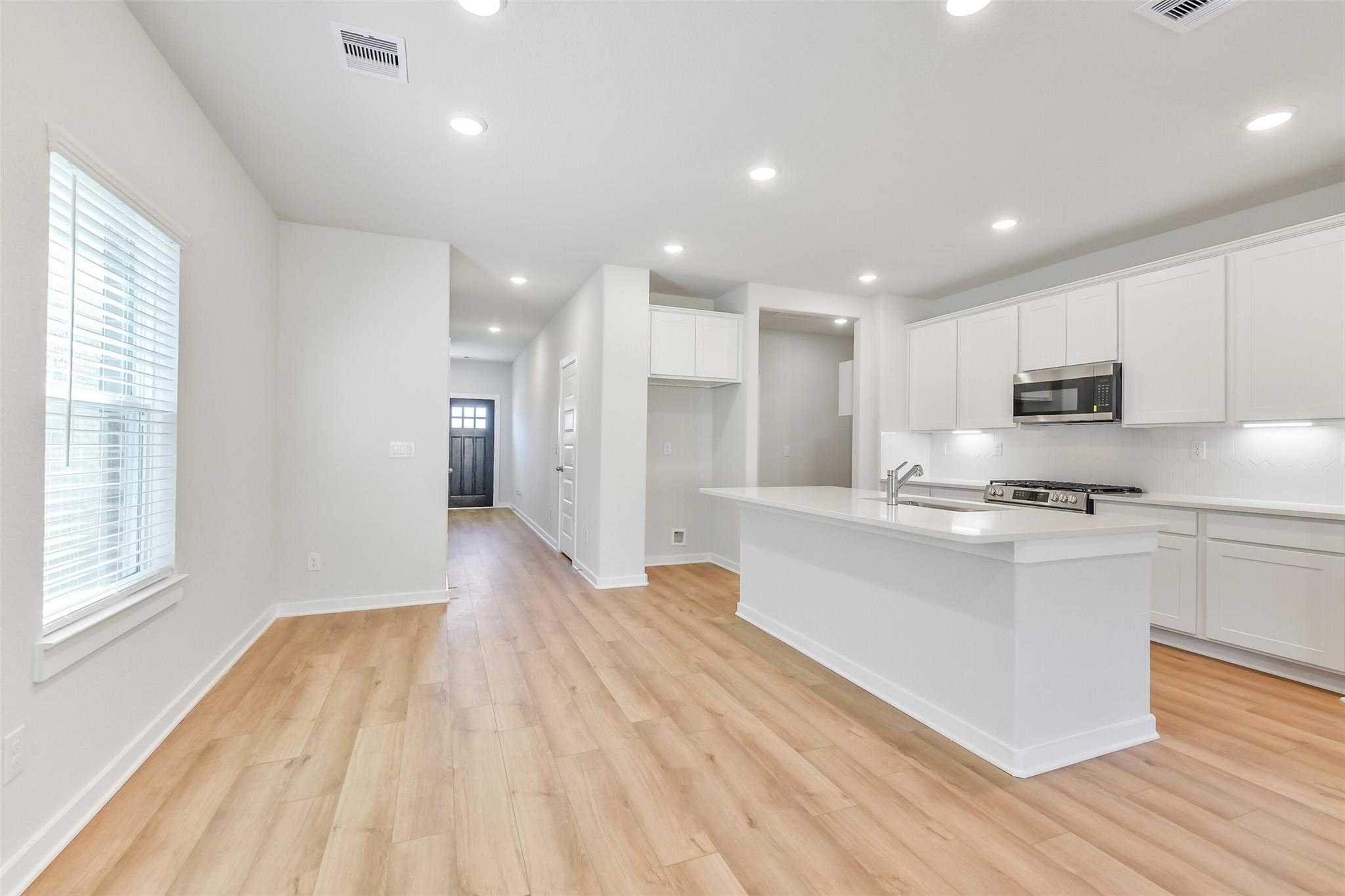 Open-concept white kitchen with large island, stainless appliances, hardwood floors, and hallway view in Davidson Homes The Blanco E, Magnolia TX