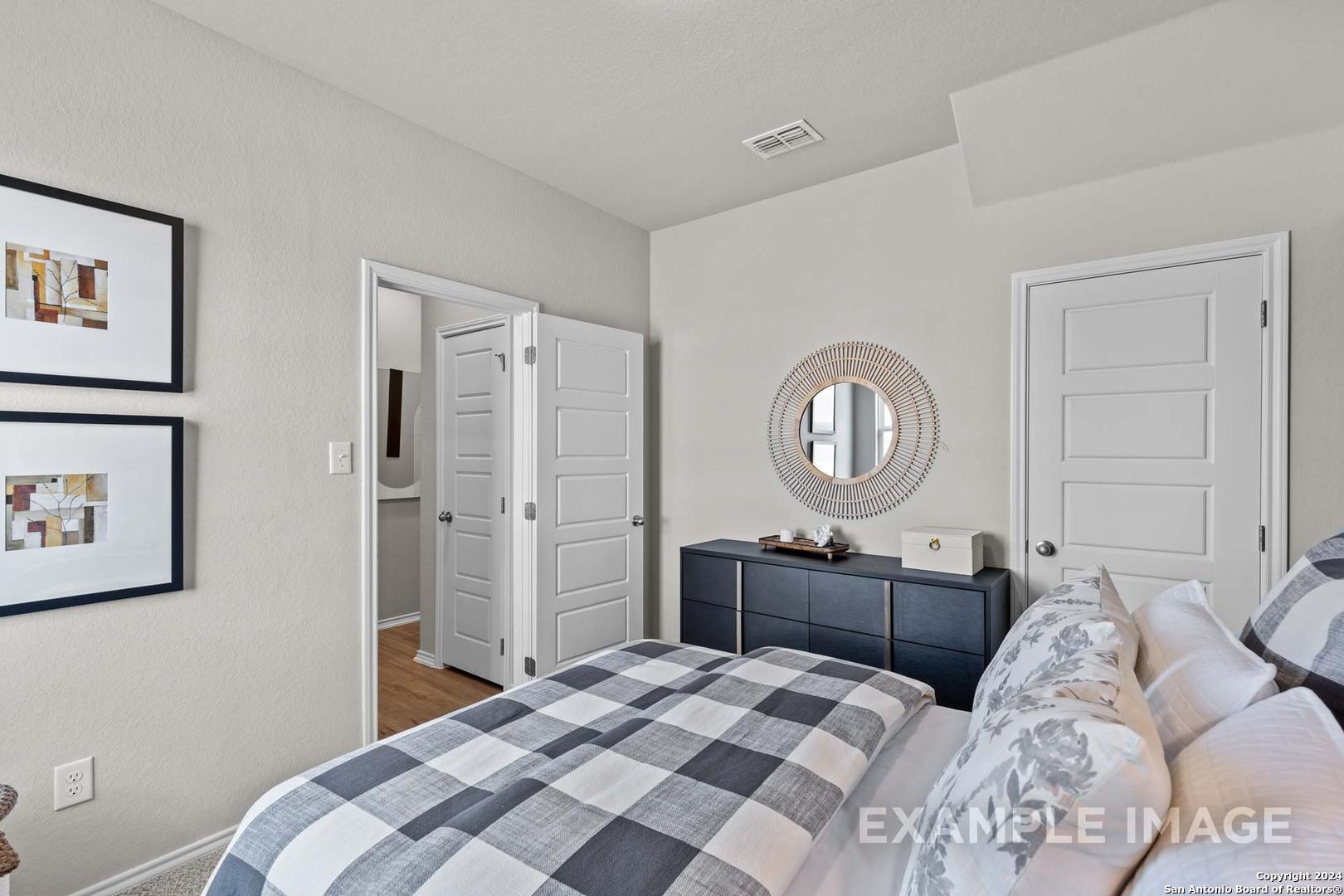 Inviting bedroom with checkered bedding, wooden dresser, round mirror, and bath access in Davidson Homes The Sabine B, San Antonio TX