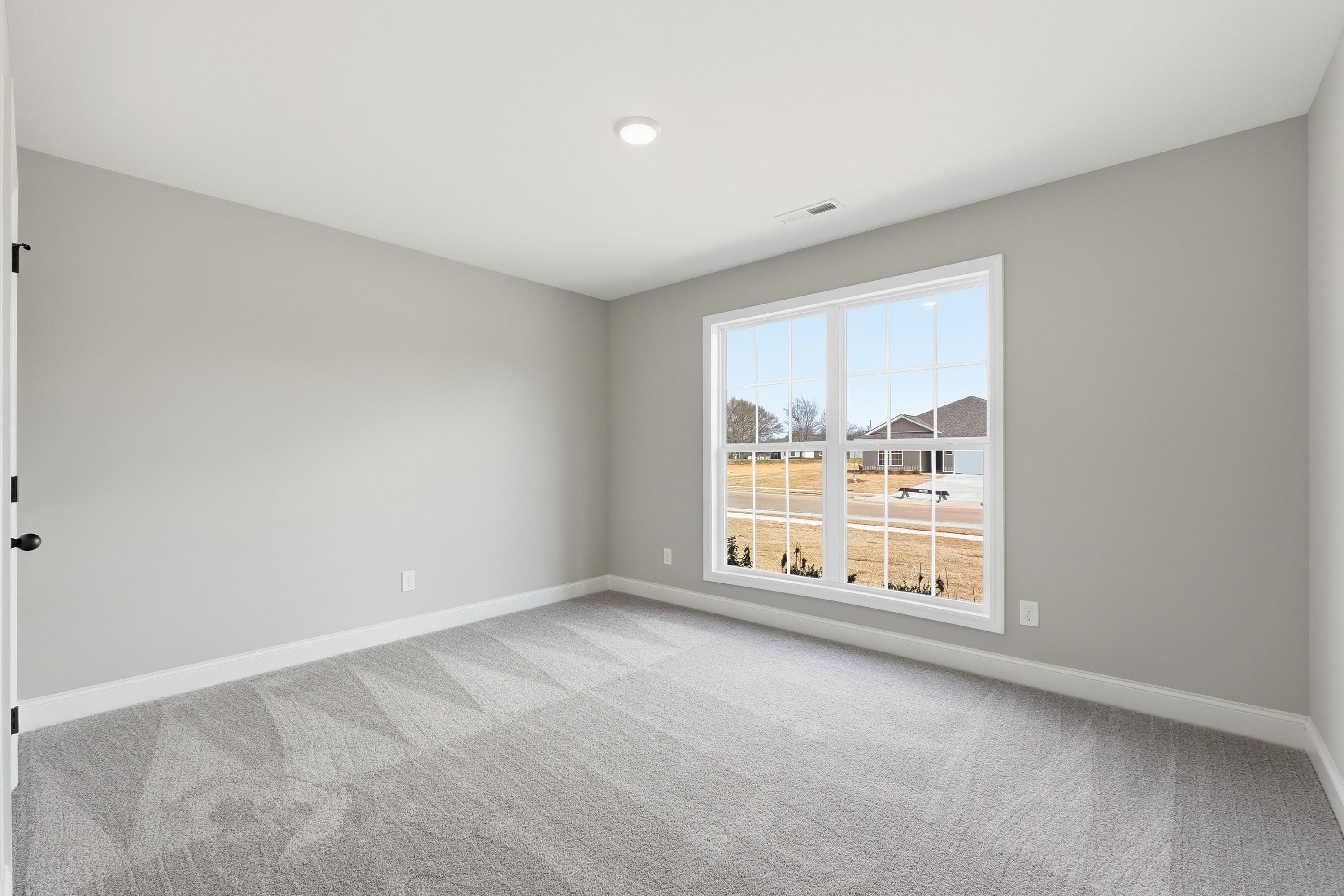 Spacious empty bedroom in The Daphne V with gray walls, carpet, and large window overlooking rural field in Meridianville
