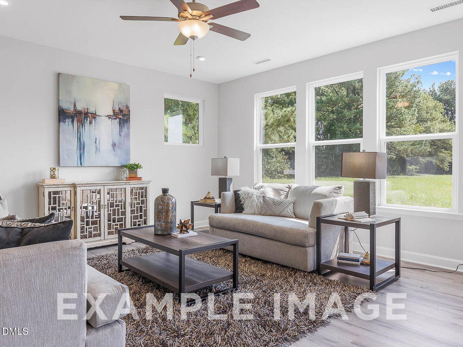 Bright modern living room with ceiling fan, large windows overlooking trees, gray sofa, and abstract art in Davidson Homes Chestnut B, Lillington, NC