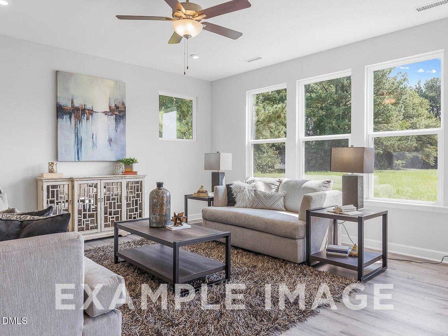 Bright modern living room with ceiling fan, large windows overlooking trees, gray sofa, and abstract art in Davidson Homes Chestnut B, Lillington, NC