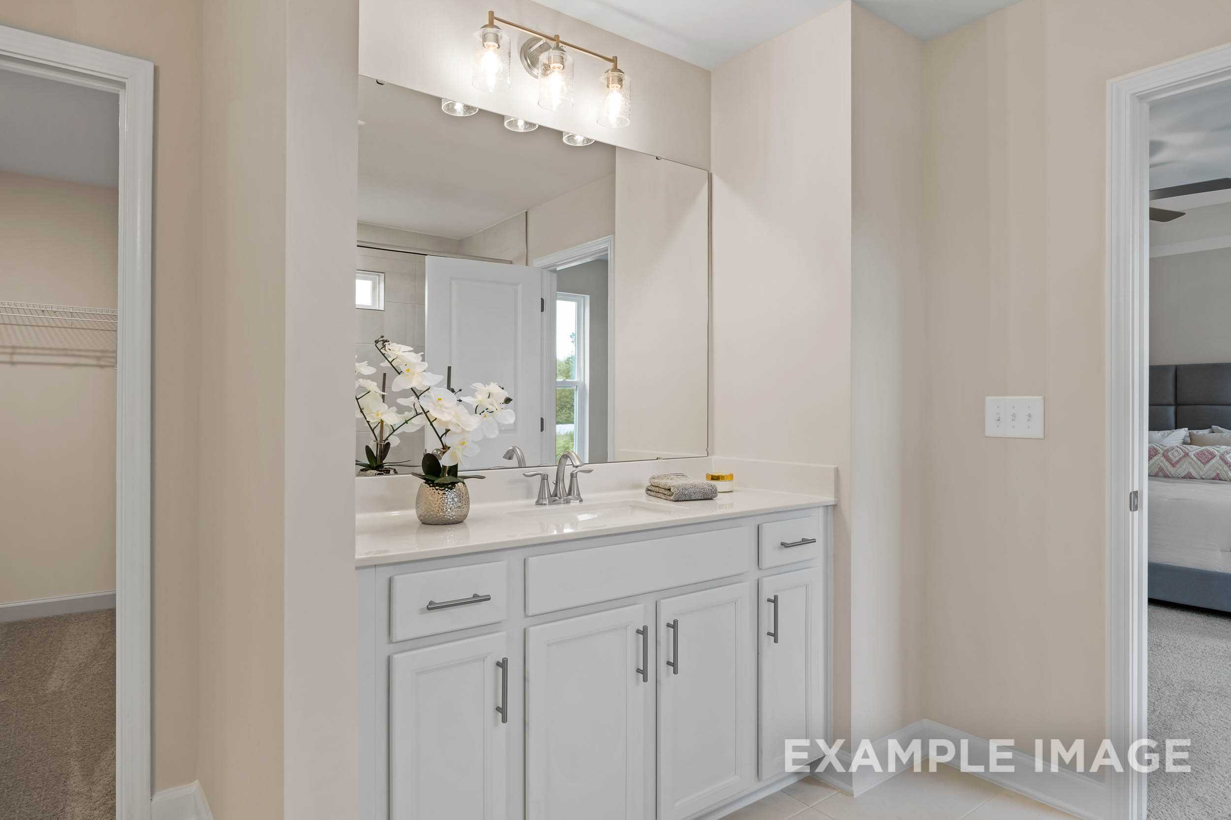 Spacious master bathroom in The Preston B with white double-sink vanity, lighted mirror, orchids, and adjacent bedroom view