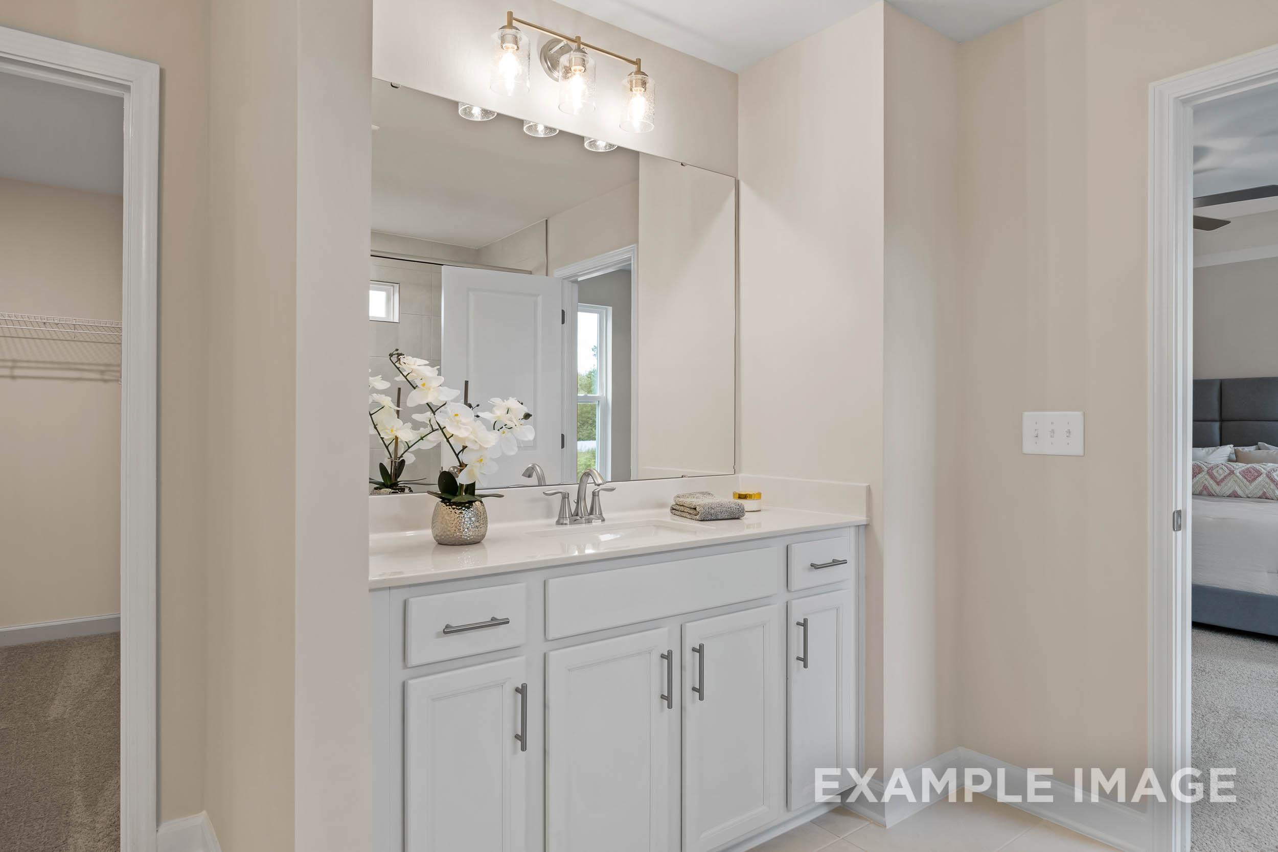 Spacious master bathroom in The Preston B with white double-sink vanity, lighted mirror, orchids, and adjacent bedroom view