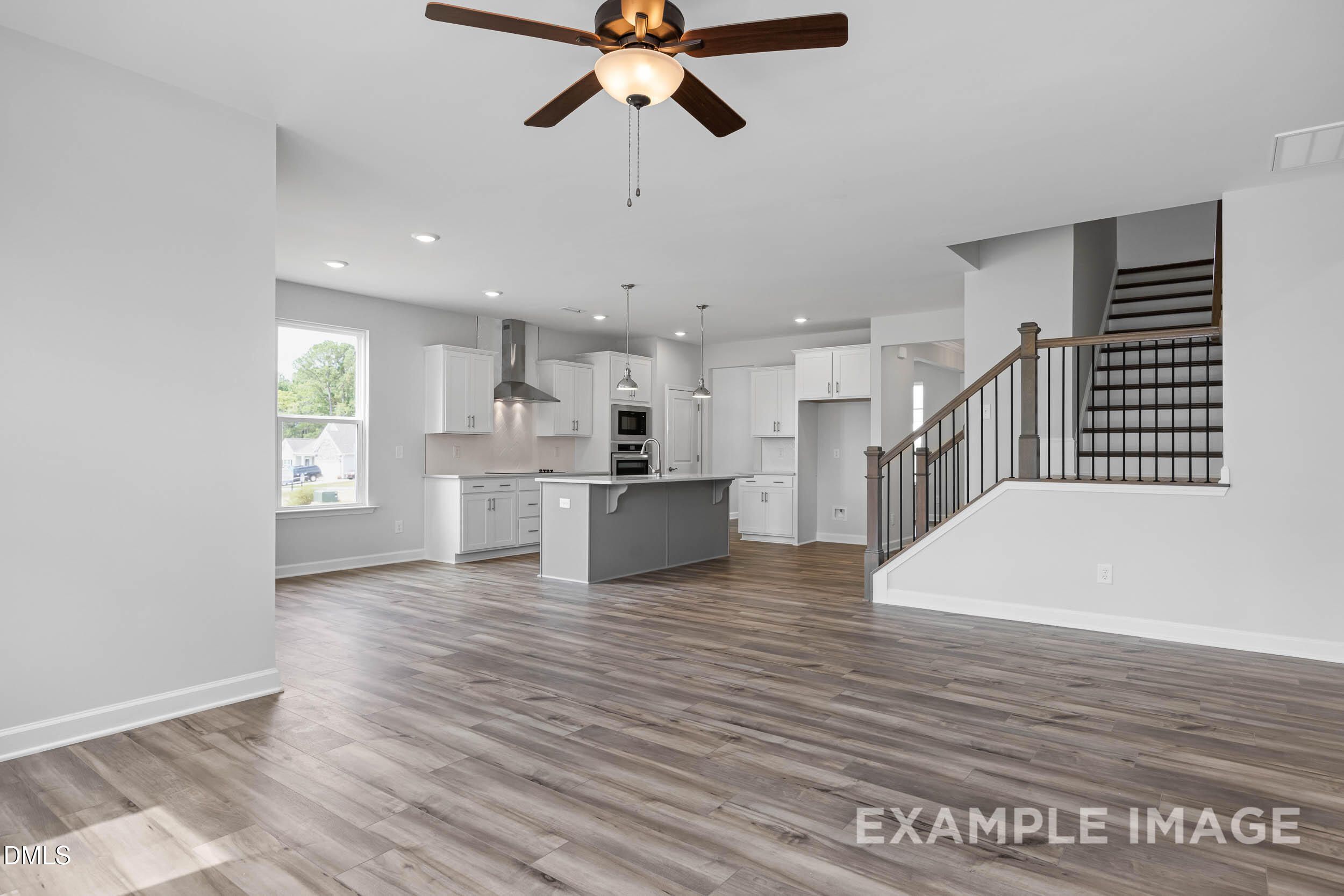 Spacious open-concept living room with hardwood floors, modern gray kitchen island, white cabinets, ceiling fan, and oak staircase in Davidson Homes The Ash B, Lillington, NC