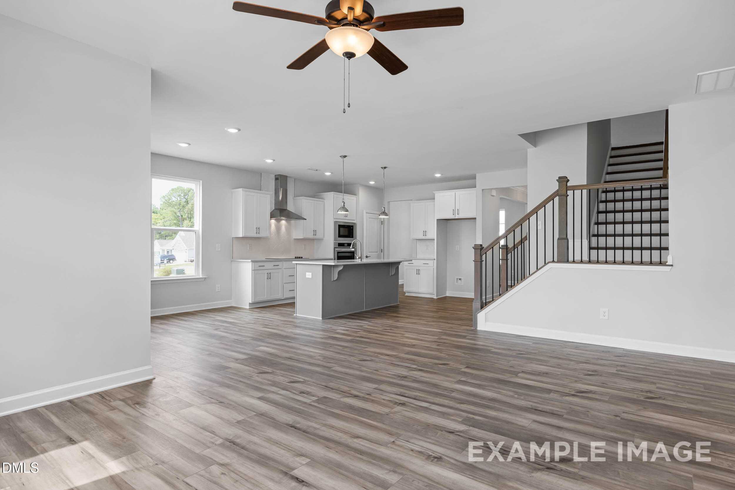 Spacious open-concept living room with hardwood floors, modern gray kitchen island, white cabinets, ceiling fan, and oak staircase in Davidson Homes The Ash B, Lillington, NC