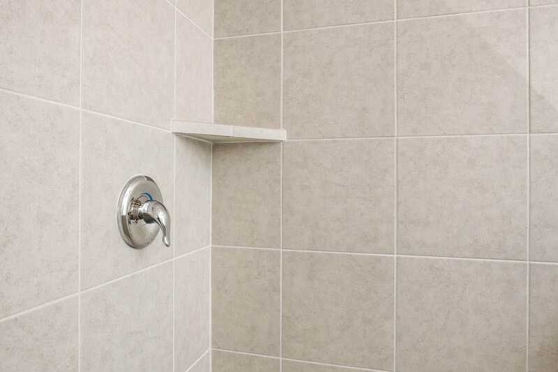 Beige tiled shower enclosure in The Gordon B master suite, featuring chrome showerhead, faucet, built-in shelf, and pan