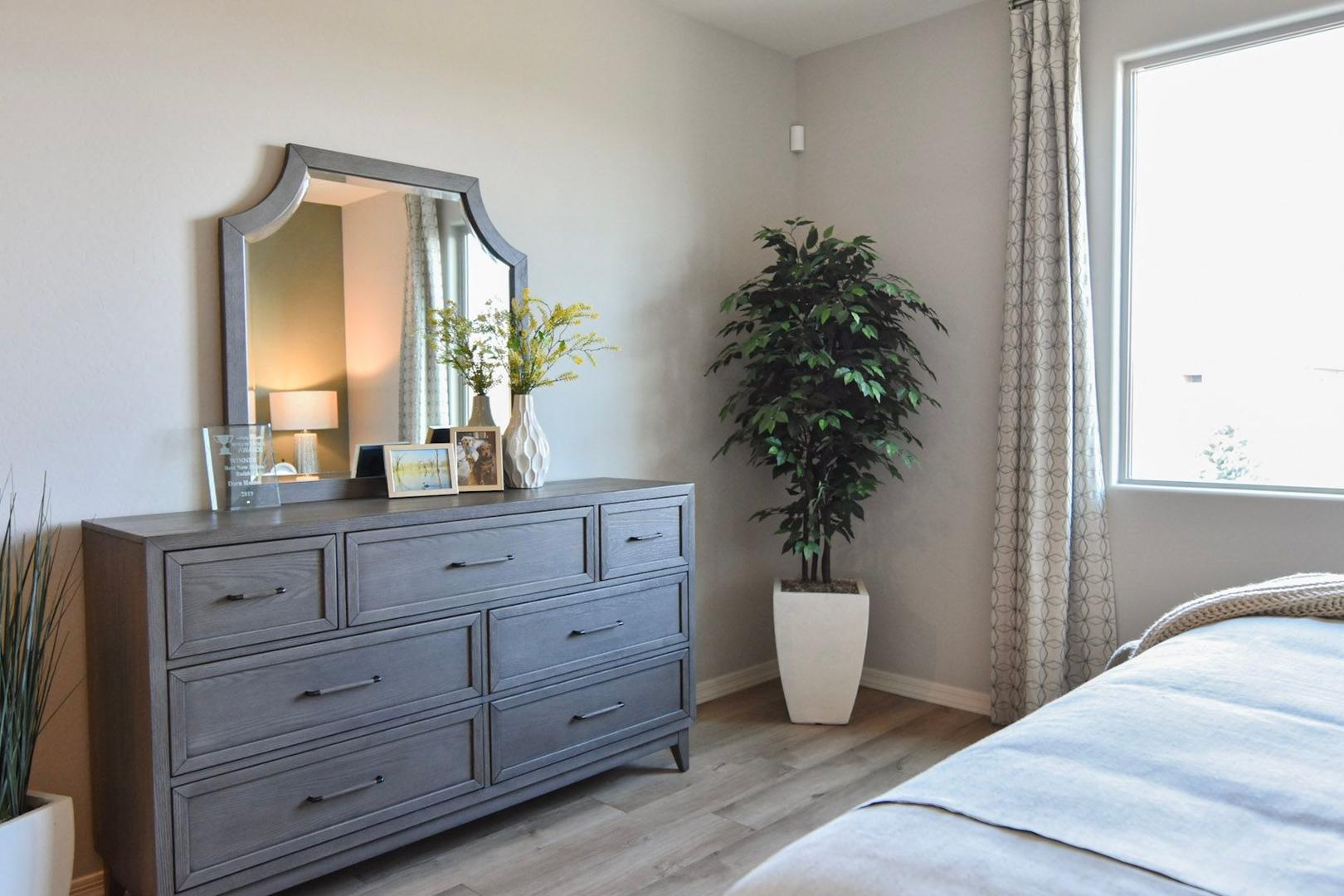 Spacious master bedroom in The Harmony B with gray dresser, arched mirror, potted plants, and sunlit window