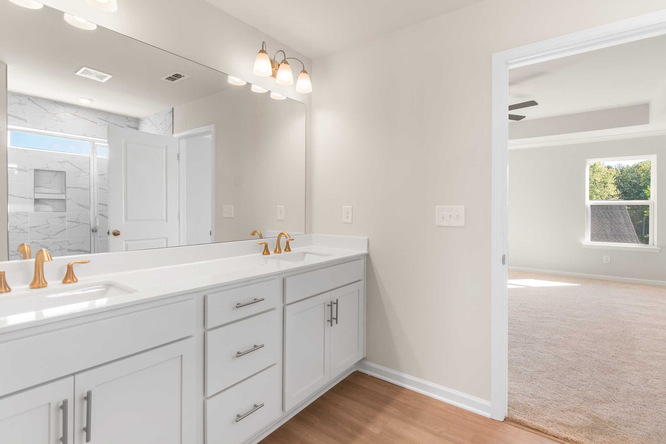Spacious master bathroom in The Willow B featuring double white vanity, gold faucets, large mirror, and tiled shower