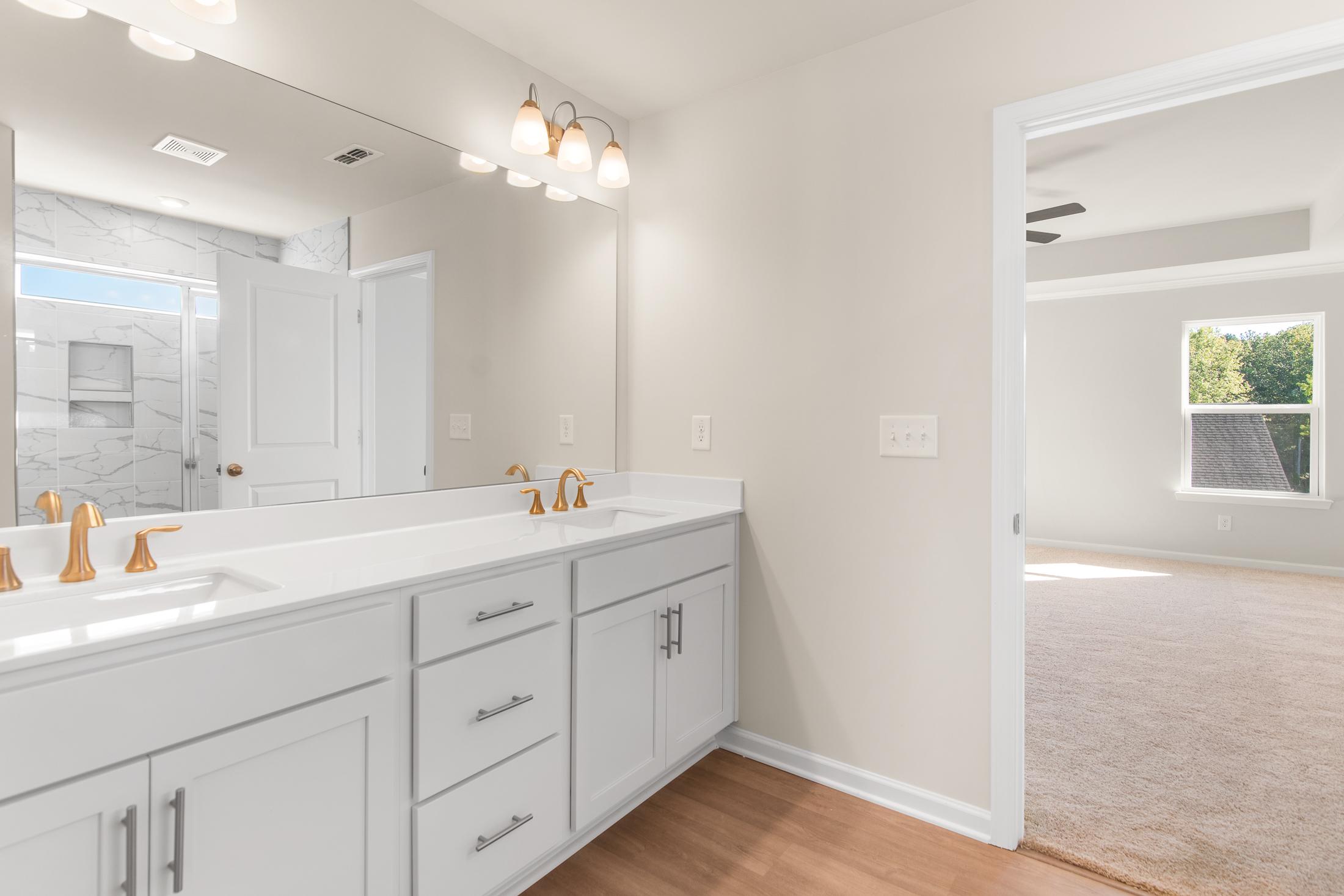 Spacious master bathroom in The Willow B featuring double white vanity, gold faucets, large mirror, and tiled shower