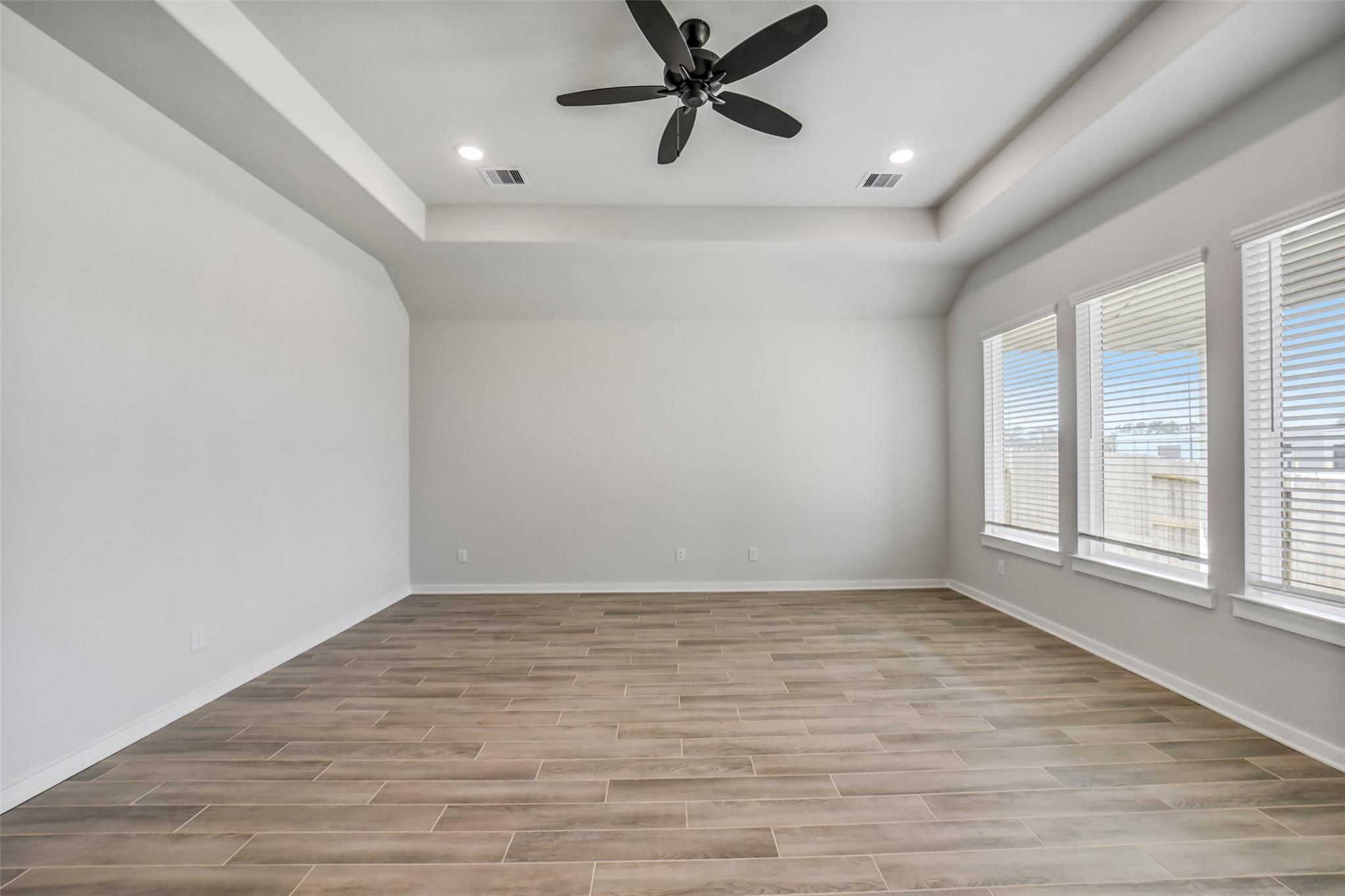 Spacious empty living room with tray ceiling, black ceiling fan, light gray walls, wood-look floors, and large windows in Davidson Homes Acadia C, Magnolia TX