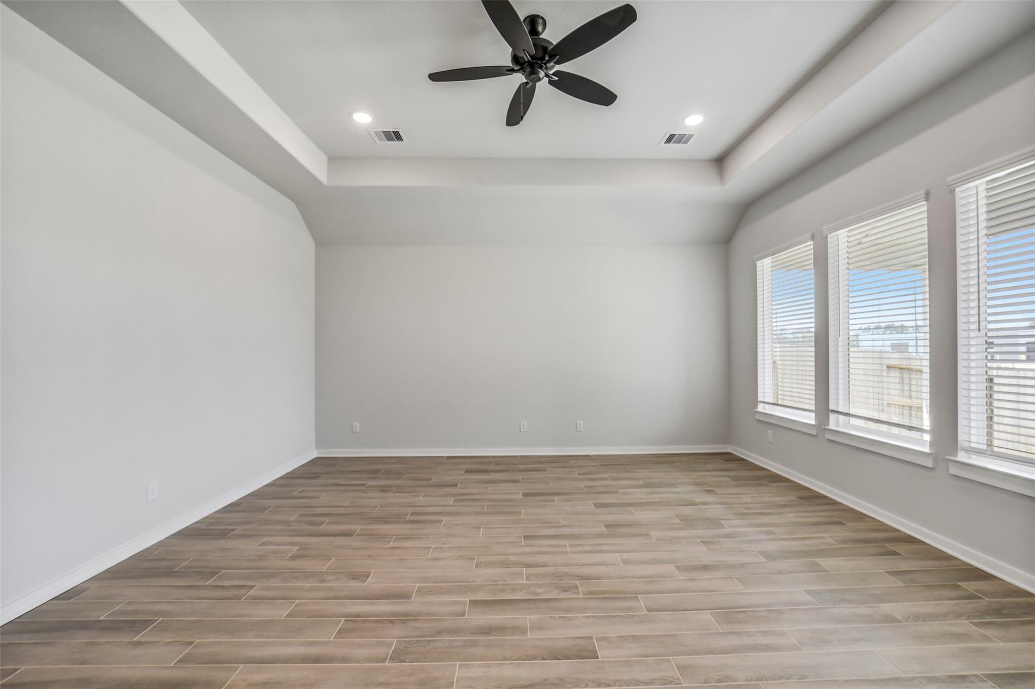 Spacious empty living room with tray ceiling, black ceiling fan, light gray walls, wood-look floors, and large windows in Davidson Homes Acadia C, Magnolia TX