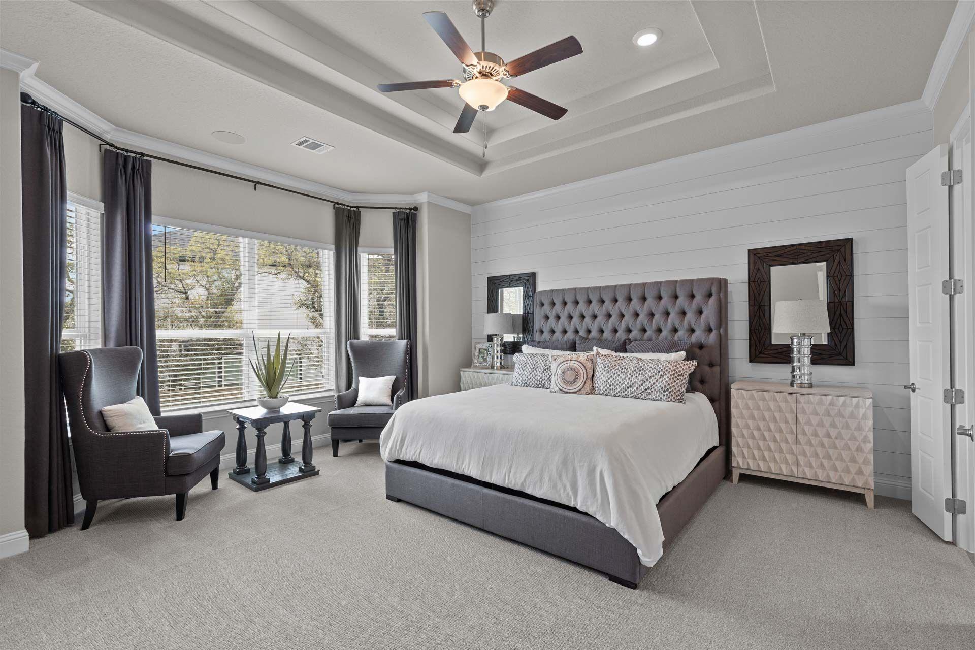 Spacious master bedroom in Ladera San Antonio with king bed, tufted gray headboard, armchairs, and bay windows