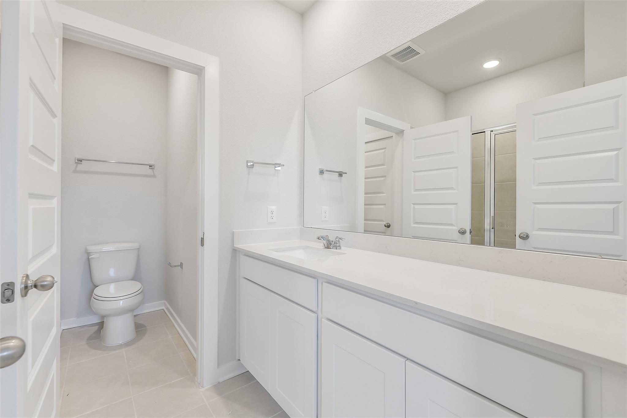 Modern white bathroom with double vanity, large mirror, toilet, and adjacent shower in Davidson Homes The Colorado F, Cleveland, Texas