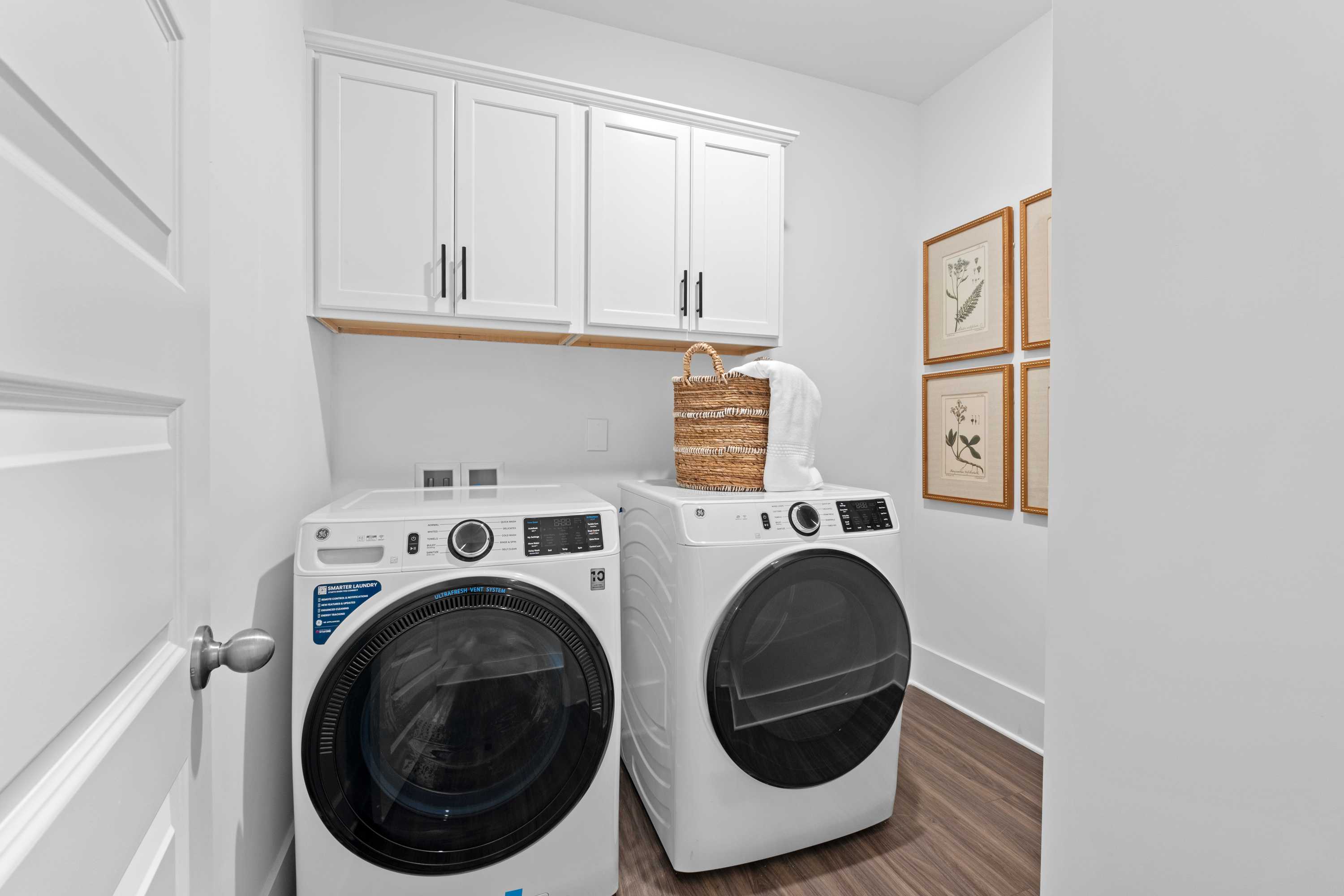 Bright laundry room at Ivy Hills in Toney Alabama with white washer dryer, overhead cabinets, wicker basket, and hardwood floors