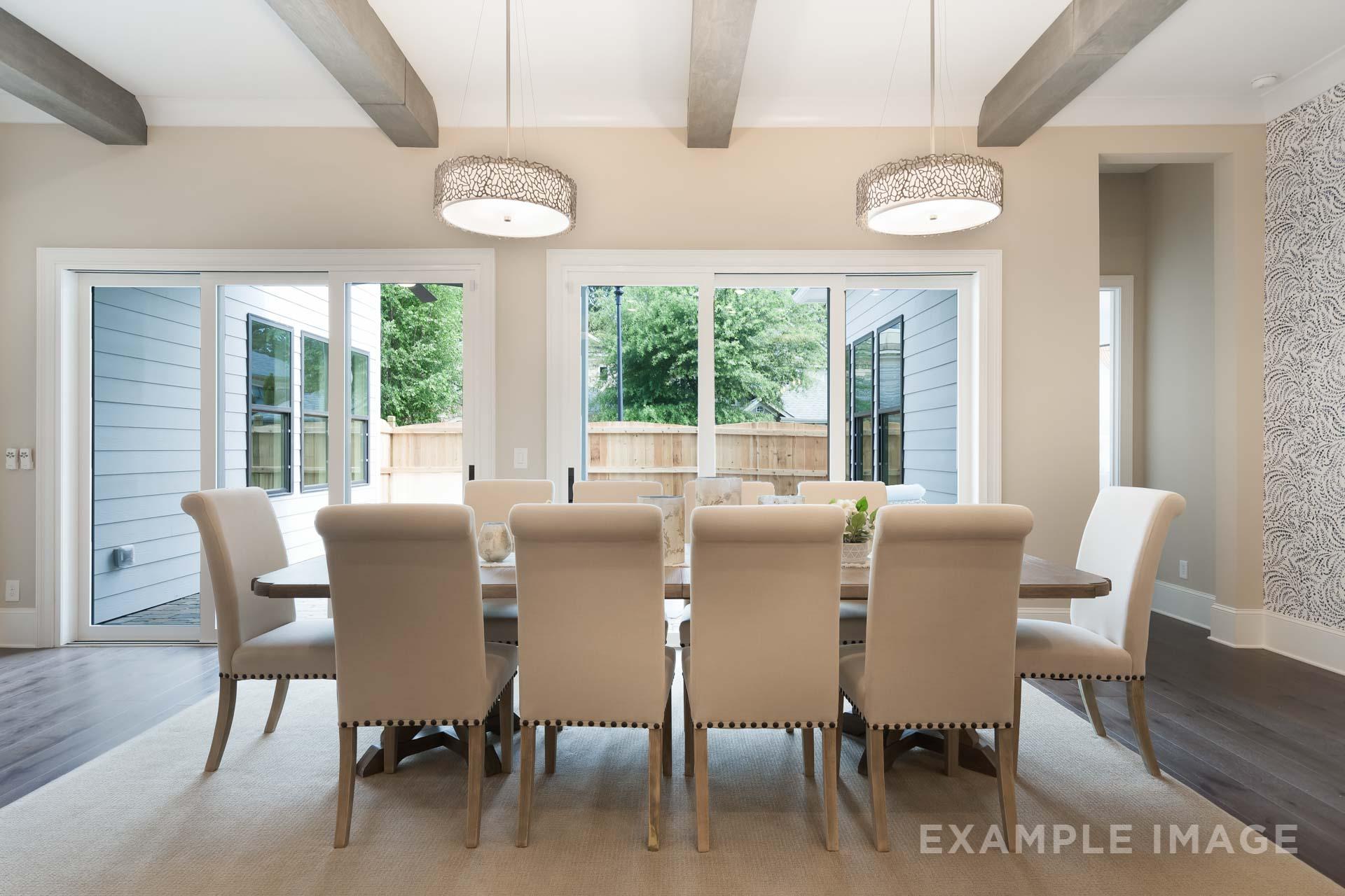Spacious dining room in The Seaside C featuring long wooden table, beige upholstered chairs, beamed ceiling, and French doors to wooded backyard
