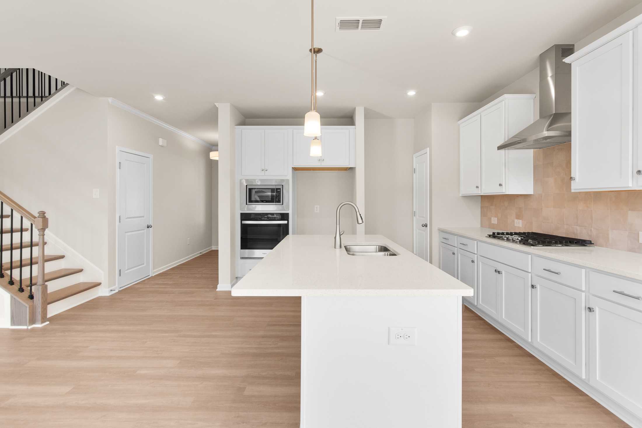 Spacious open-concept kitchen in The Willow B showcasing white cabinets, quartz island, stainless appliances, and adjacent staircase at Wehunt Meadows, Hoschton GA