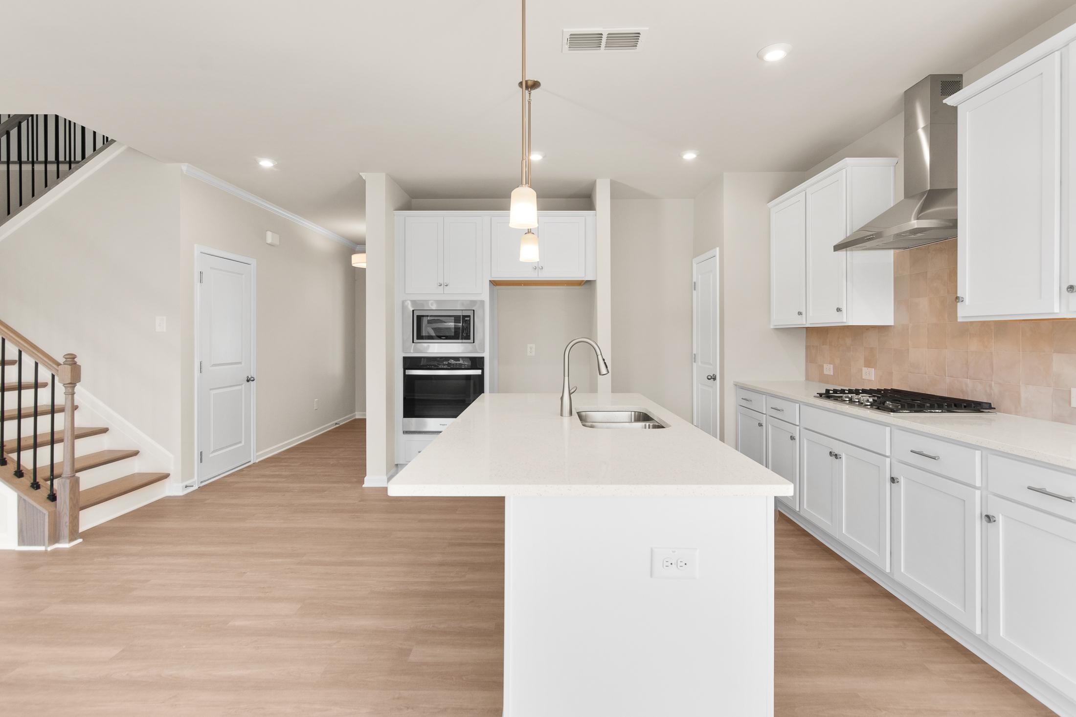 Spacious open-concept kitchen in The Willow B showcasing white cabinets, quartz island, stainless appliances, and adjacent staircase at Wehunt Meadows, Hoschton GA