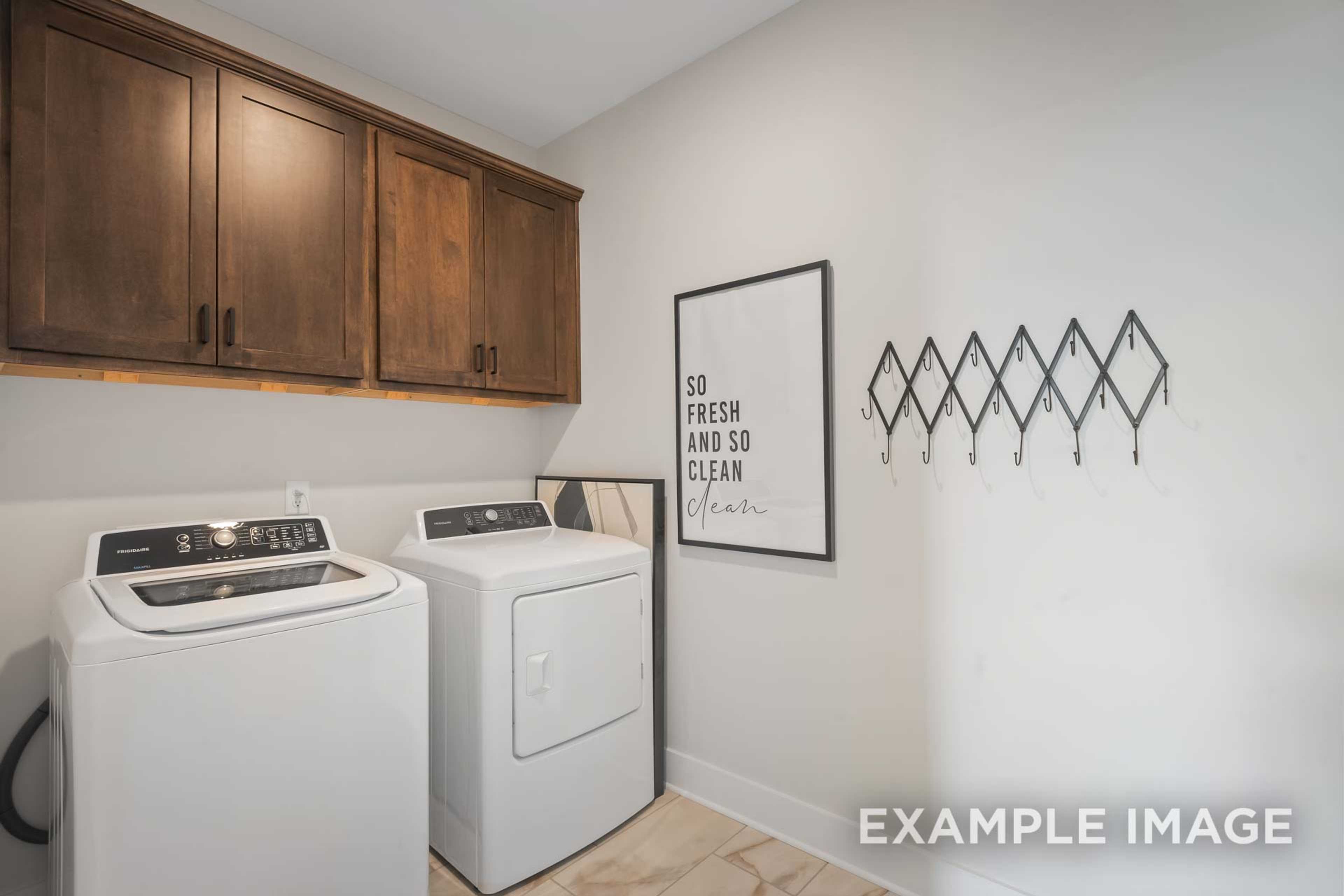 Spacious laundry room in The Ridgeport B with white washer dryer, wooden upper cabinets, and "So fresh and so clean" wall art