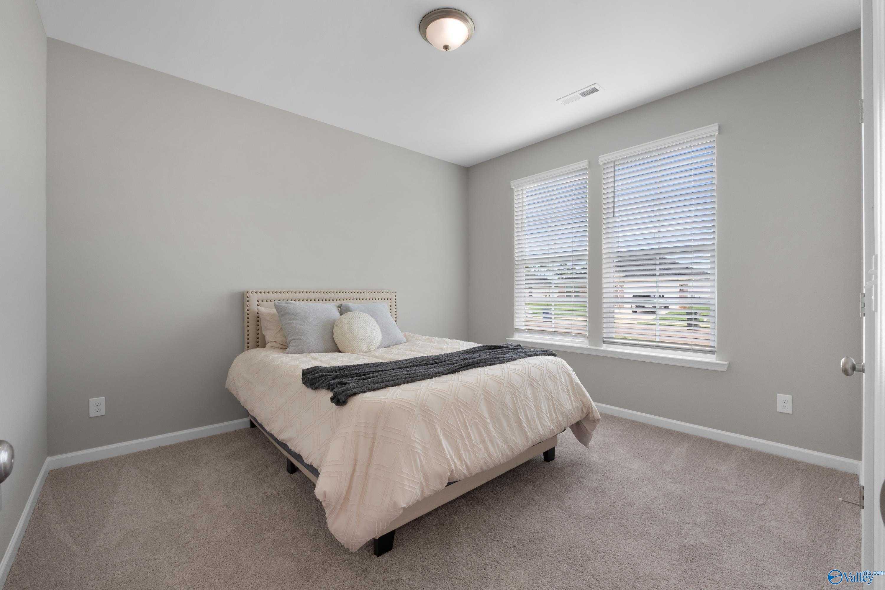 Neutral-toned secondary bedroom with rattan headboard bed, beige bedding, and double windows in Evermore Homes The Noble, Madison Alabama