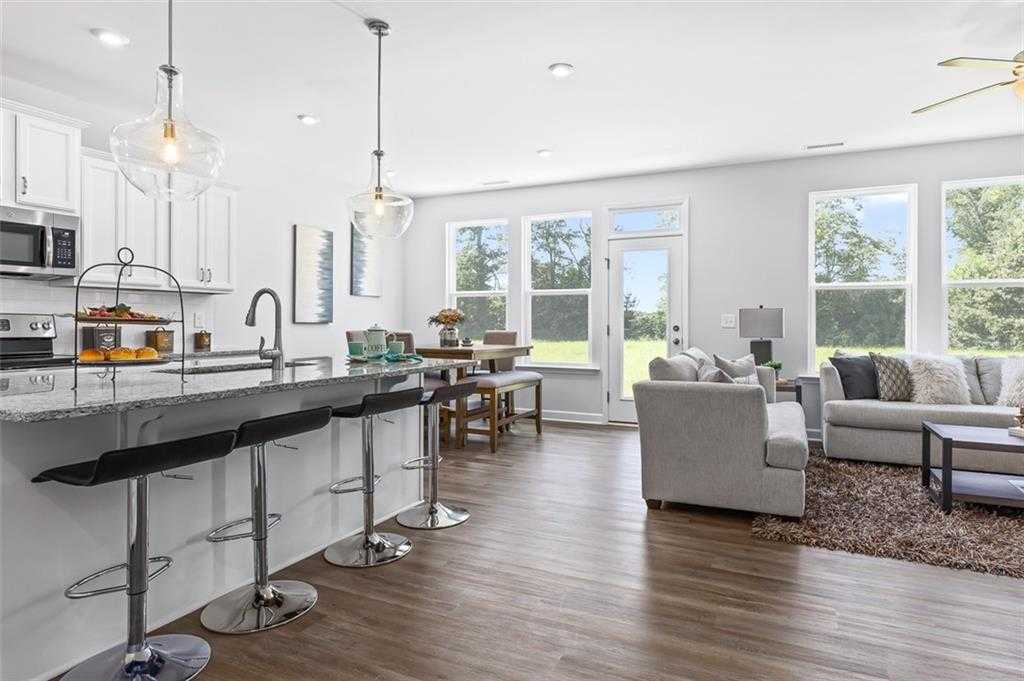 Modern open-concept kitchen with white cabinets, granite island, bar stools, and adjacent living room in Davidson Homes The Willow E at Wehunt Meadows, Hoschton, GA