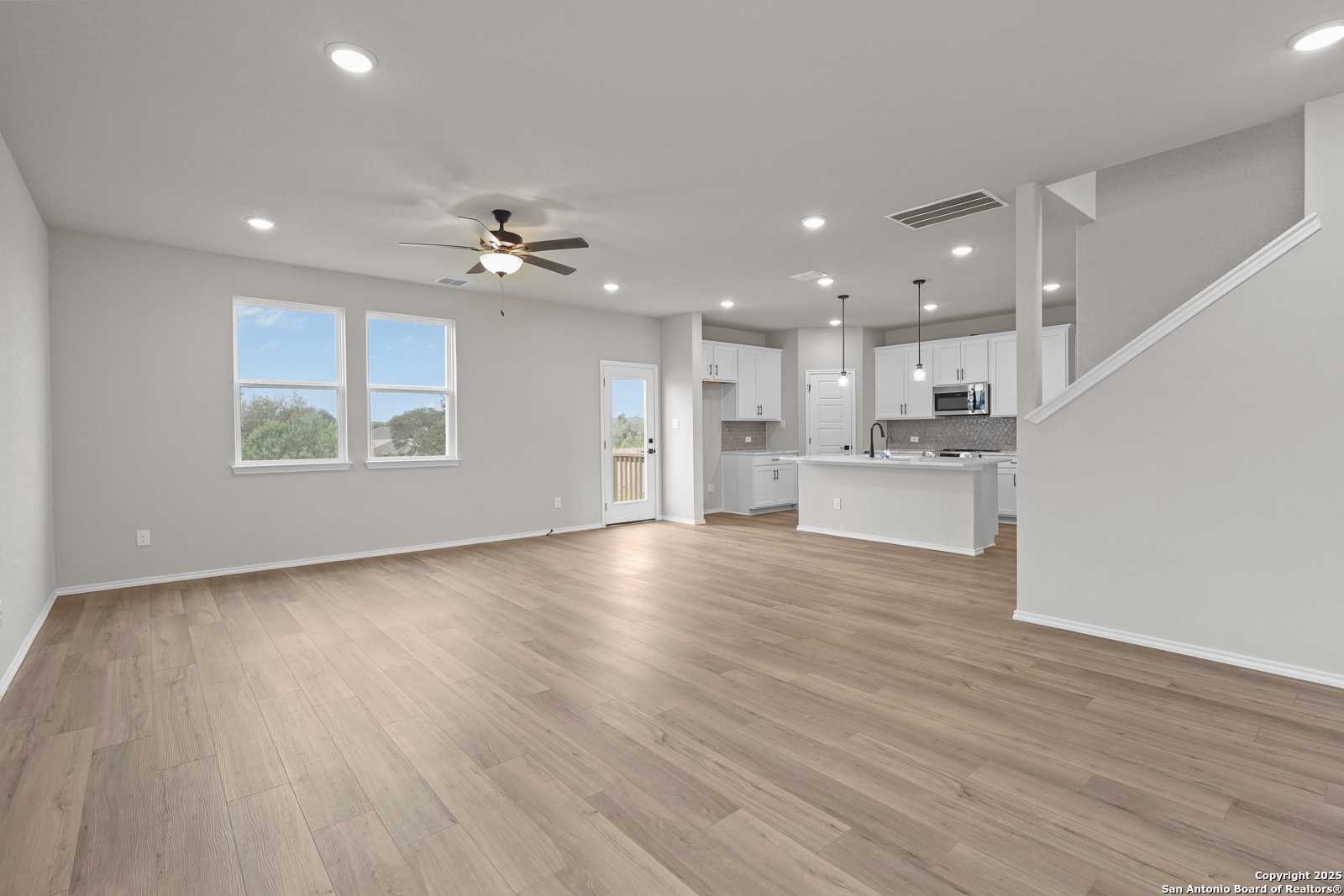Open-concept living and kitchen with hardwood floors, white cabinets, ceiling fan, and large windows in Davidson Homes The Murray J, San Antonio