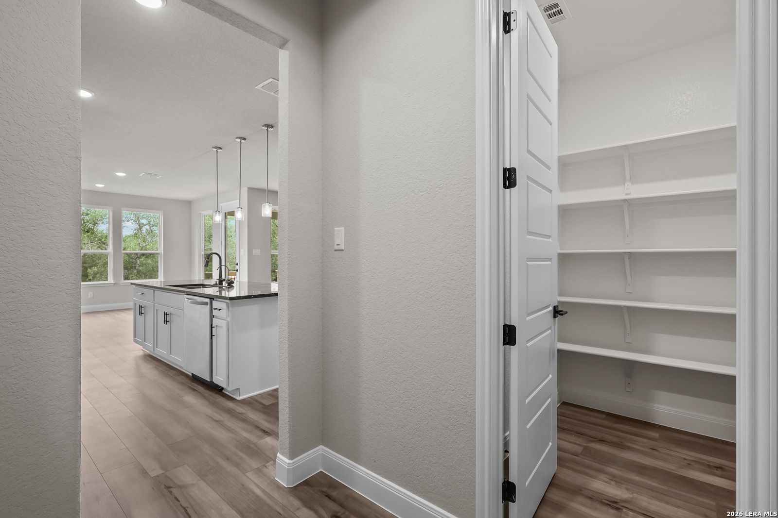 Spacious walk-in pantry with white shelving adjacent to open kitchen island, sink, and pendant lights in Davidson Homes The Belmont H, Castroville TX