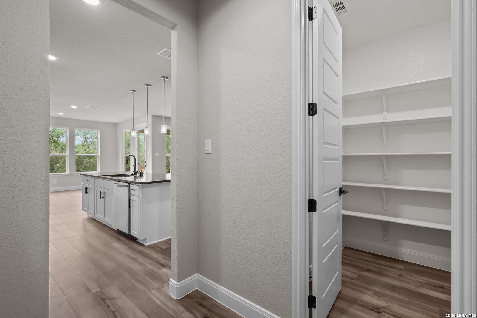 Spacious walk-in pantry with white shelving adjacent to open kitchen island, sink, and pendant lights in Davidson Homes The Belmont H, Castroville TX