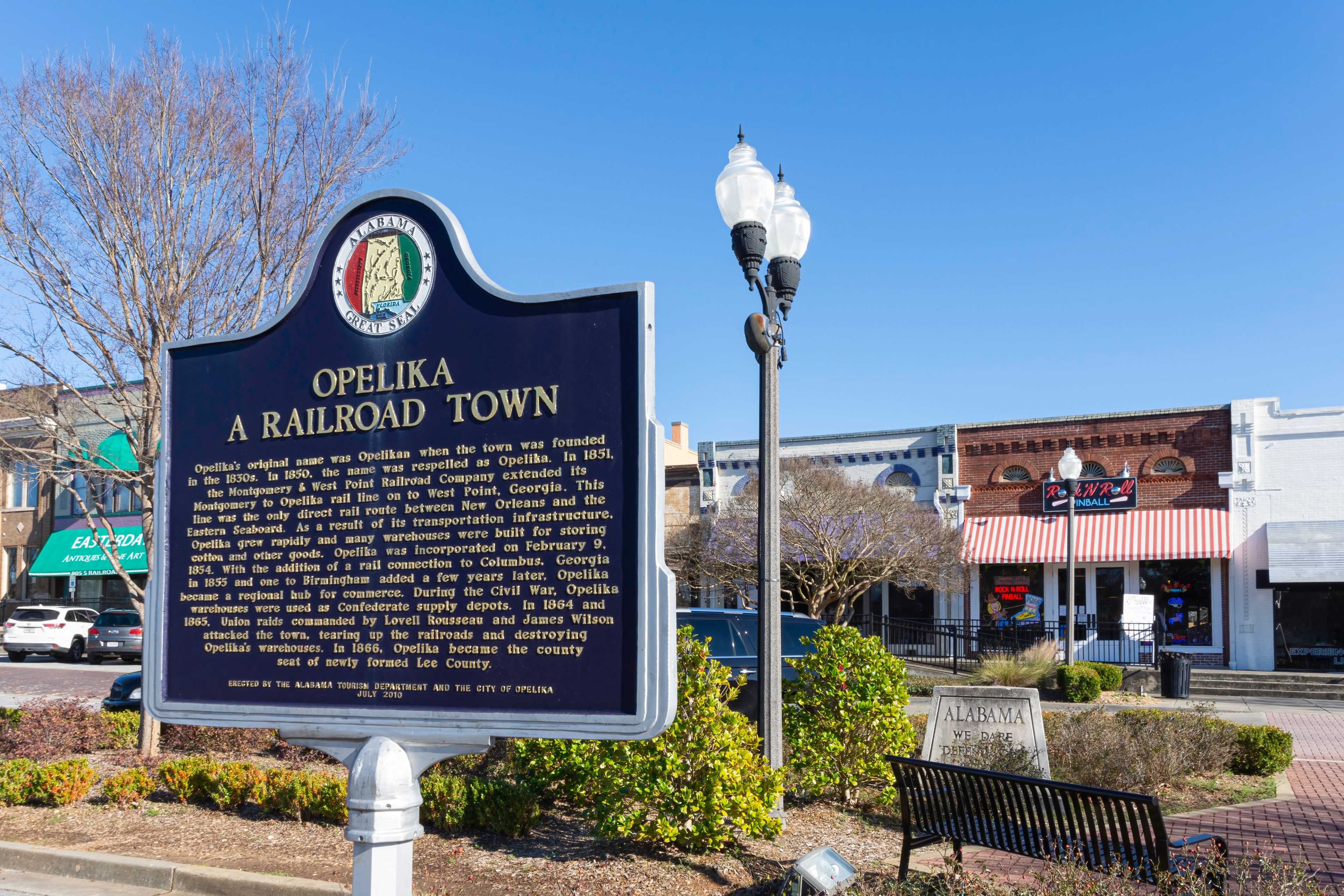 Historical marker on Opelika's railroad town founding in downtown Opelika Alabama with historic buildings, lamps, and park bench