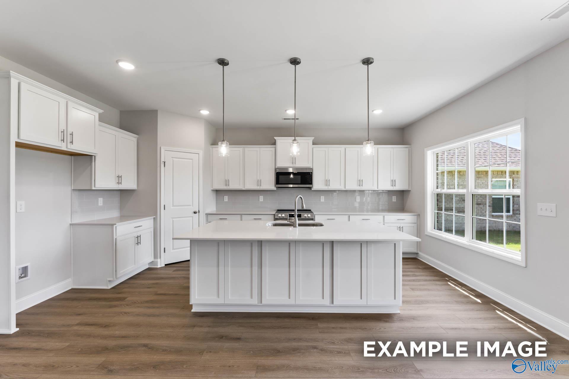 Modern white kitchen with large island, stainless appliances, pendant lights, and hardwood floors in The Madison A, Meridianville, Alabama