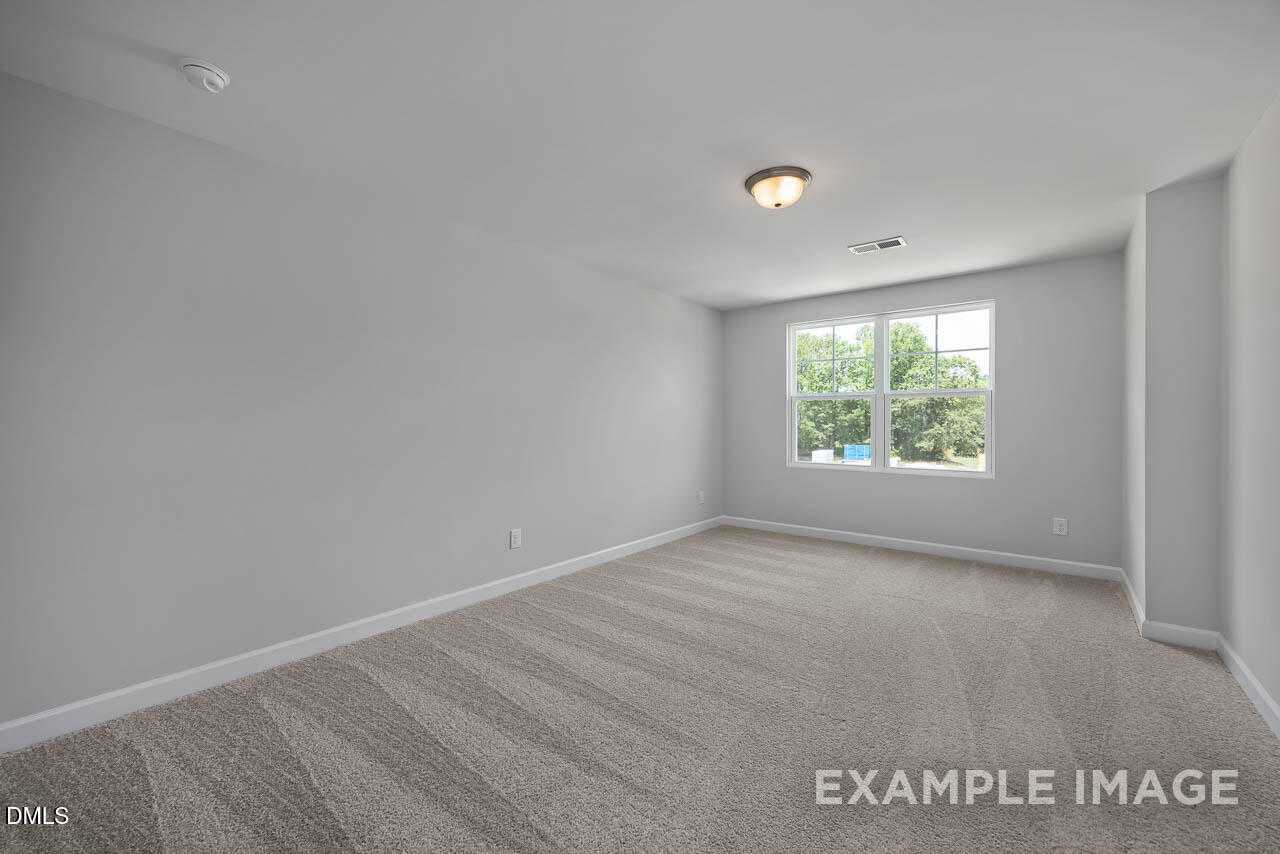 Bright empty bedroom with light gray walls, beige carpet, and large window overlooking trees in Davidson Homes The Warren, Fuquay-Varina