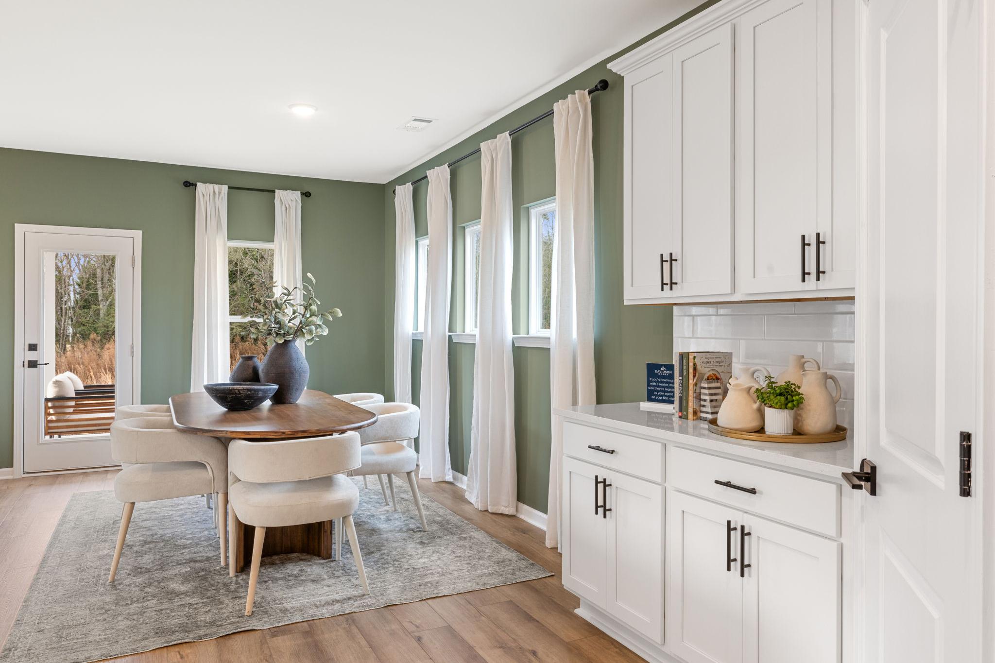 Spacious dining area with sage green walls, wooden table, white cabinets, and potted plants at Links Crossing in Auburn, Alabama