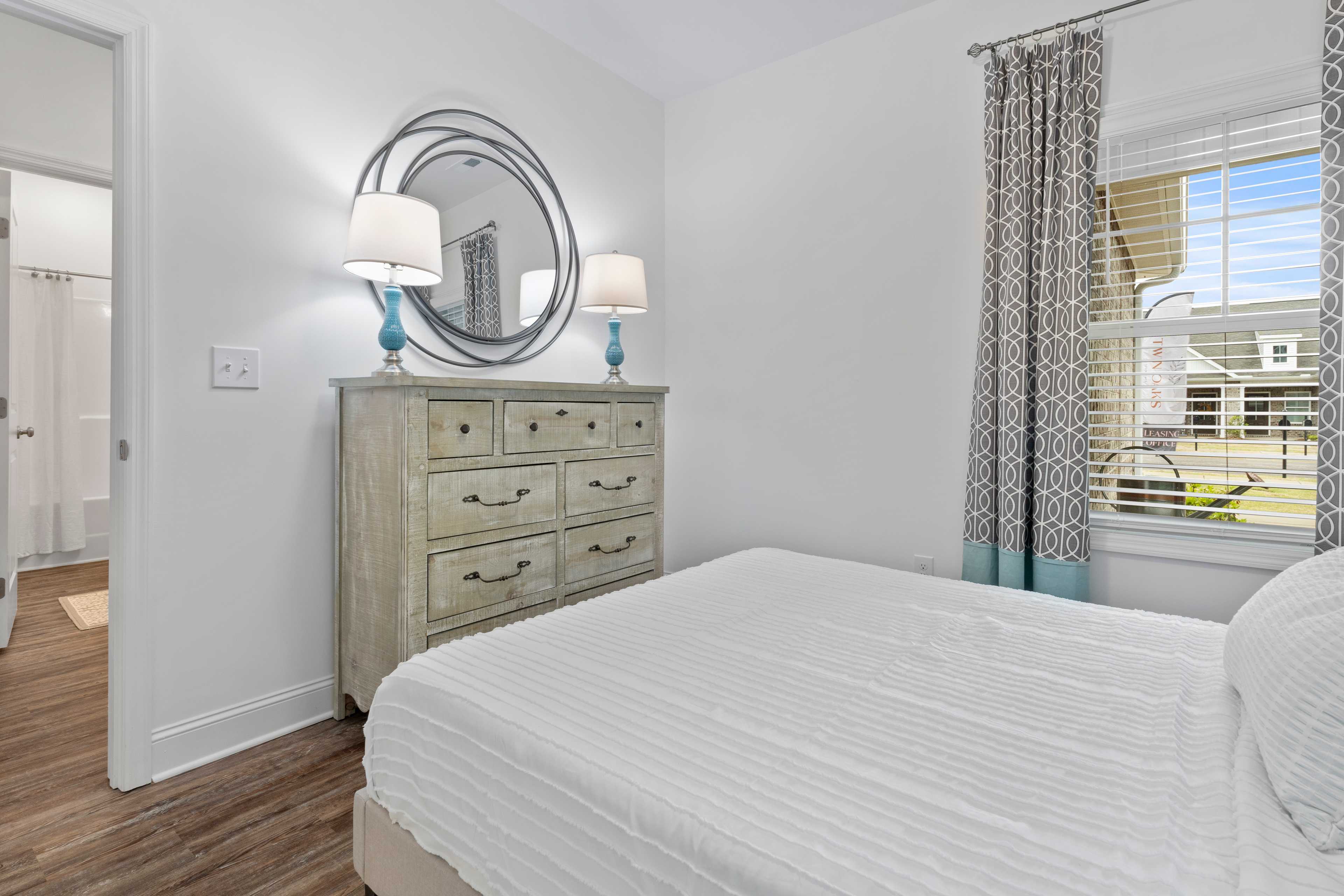 Spacious bedroom at The Retreat at Hollon Meadow in Decatur Alabama featuring white bed, rustic dresser, round mirror, and hardwood floors